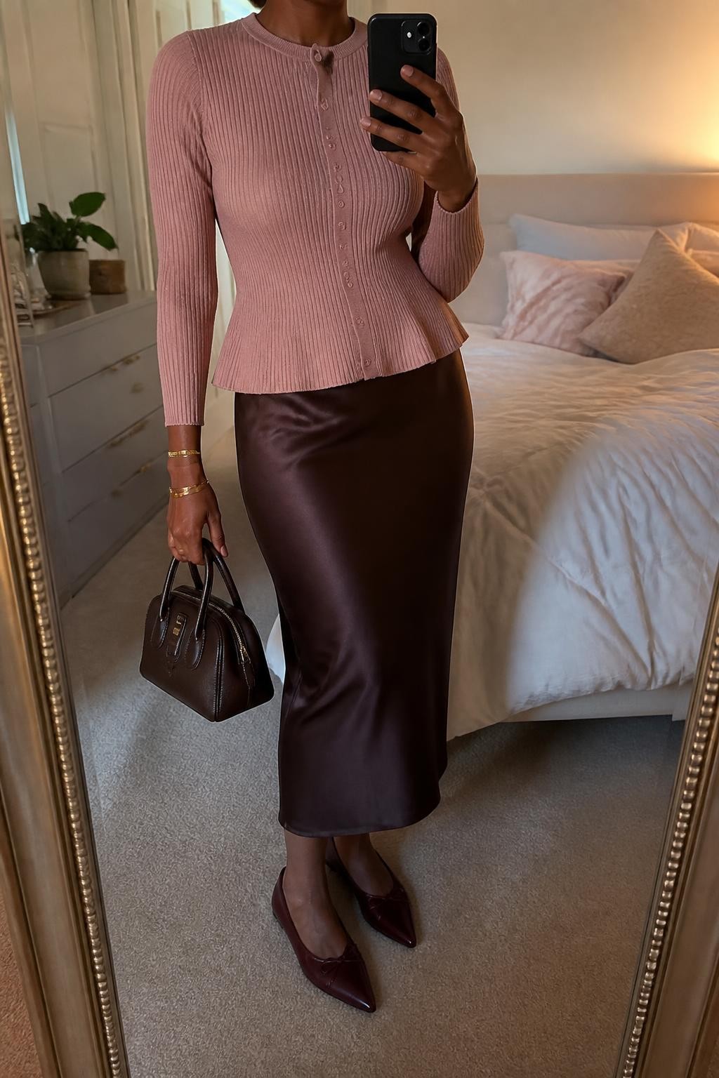 mirror selfie of a Black woman wearing a fully buttoned dusty pink ribbed peplum cardigan, a chocolate brown satin midi slip skirt, burgundy pointed-toe ballet flats, a chocolate structured mini handbag, and a slim gold cuff bracelet, clearly showing the exact clothing items, colors, fabrics, shoes, bag, and bracelet listed in the Outfit Pieces section, holding a phone in front of a mirror, cropped at NECK so the face does not show, relaxed feminine selfie pose in a feminine-themed bedroom, evening golden hour light, with soft pillows on an unmade bed and a small plant on a dresser, realistic ribbed knit, satin, leather-look, and metal textures, latest iPhone photo quality