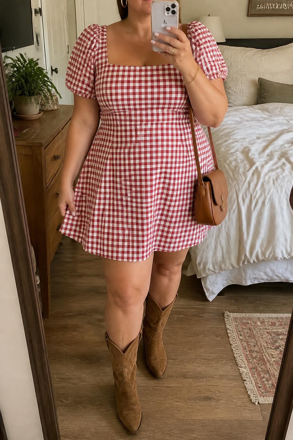 Mirror selfie of a plus size woman wearing a red and white gingham A-line mini dress with short puff sleeves, a square neckline, and a softly fitted waist, tan suede mid-calf Western boots, a small tan leather saddle bag, and delicate gold hoop earrings, clearly showing the exact dress pattern, colors, fabrics, sleeve shape, footwear, and accessories listed, holding a phone in front of a mirror, cropped at the neck so her face does not appear, relaxed feminine selfie pose, in a cozy bedroom mirror setup with soft indoor lighting, soft pillows on an unmade bed and a small plant on a dresser, realistic daytime country concert styling, latest iPhone photo quality.