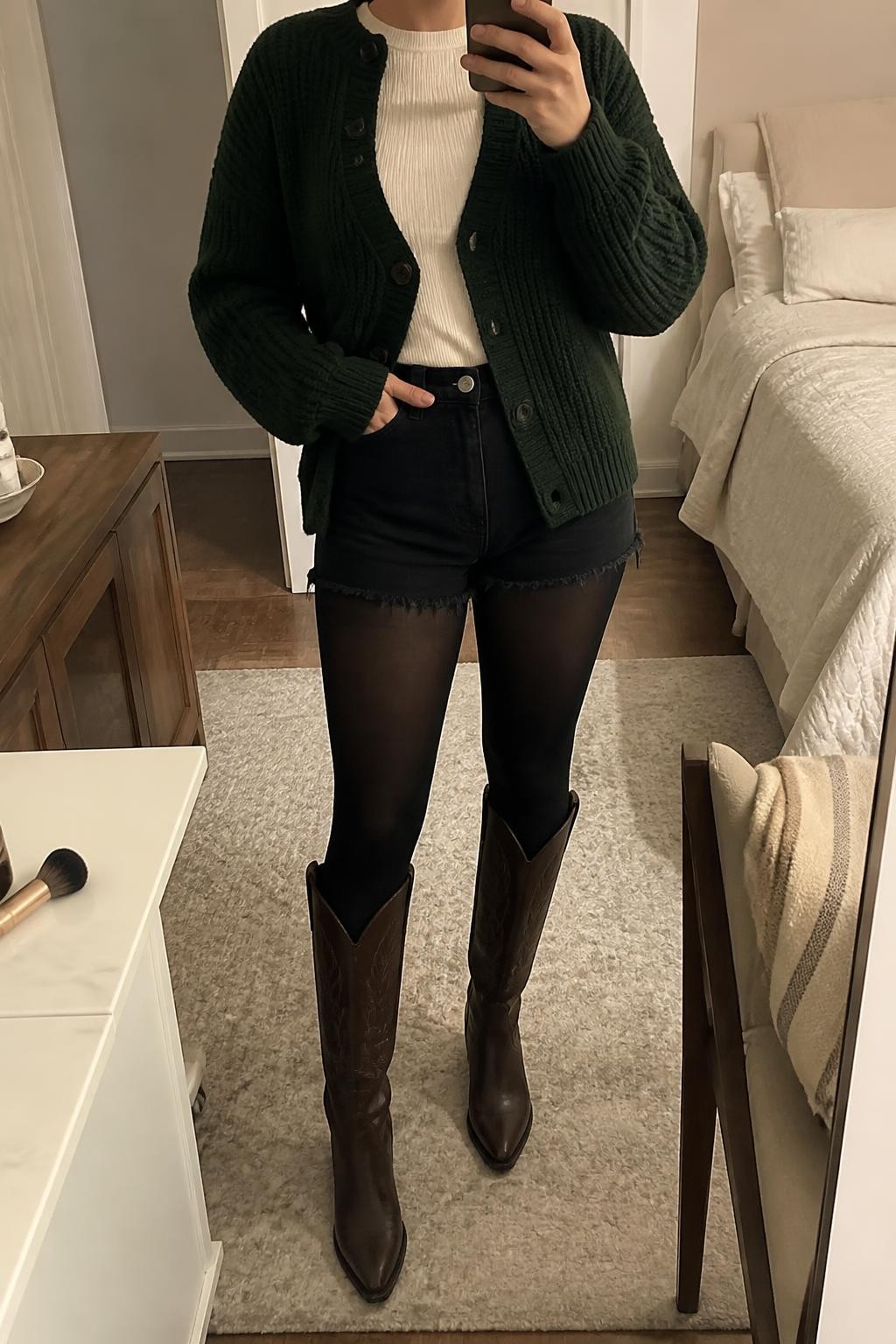 Image Prompt mirror selfie of a woman wearing a forest green chunky knit cardigan buttoned only through the middle over an ivory fitted ribbed crewneck top, black high-rise denim shorts, black opaque tights, and chocolate brown tall cowboy boots, clearly showing the exact clothing items, colors, fabrics, tights, and boots listed in the Outfit Pieces section, holding a phone in front of a mirror, cropped at NECK so the face does not show, relaxed casual selfie pose in a cozy bedroom mirror setup, soft indoor lighting, with a folded blanket on a chair and a makeup brush on a nearby counter, realistic chunky knit, ribbed knit, denim, tights, and leather textures, latest iPhone photo quality