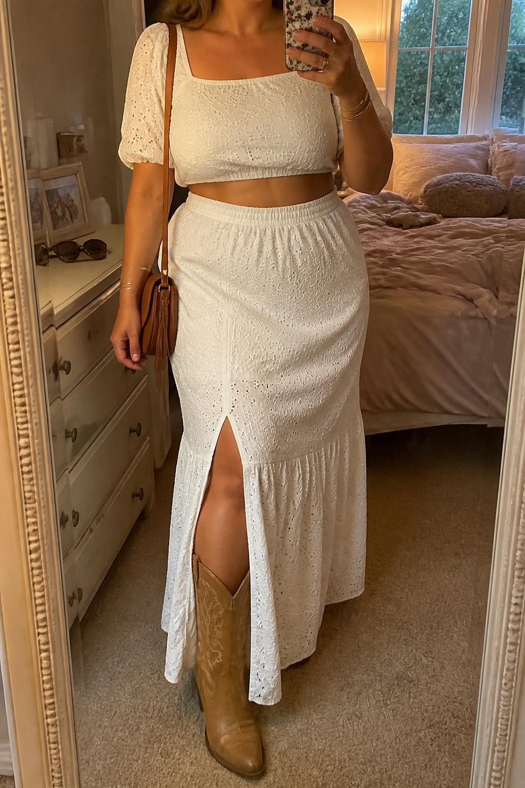 Mirror selfie of a plus size woman wearing a white cotton eyelet crop top with short puff sleeves and a square neckline, a matching white high-waisted eyelet maxi skirt with an opaque lining and side slit, tan leather mid-calf cowboy boots, and a small brown saddle bag, clearly showing the exact matching set, white eyelet texture, tan footwear, side slit, and accessories listed, holding a phone in front of a mirror, cropped at the neck so her face does not appear, relaxed feminine selfie stance, in a feminine-themed bedroom with evening golden-hour lighting, soft pillows on an unmade bed and sunglasses placed on a dresser, realistic boho festival outfit, latest iPhone photo quality.