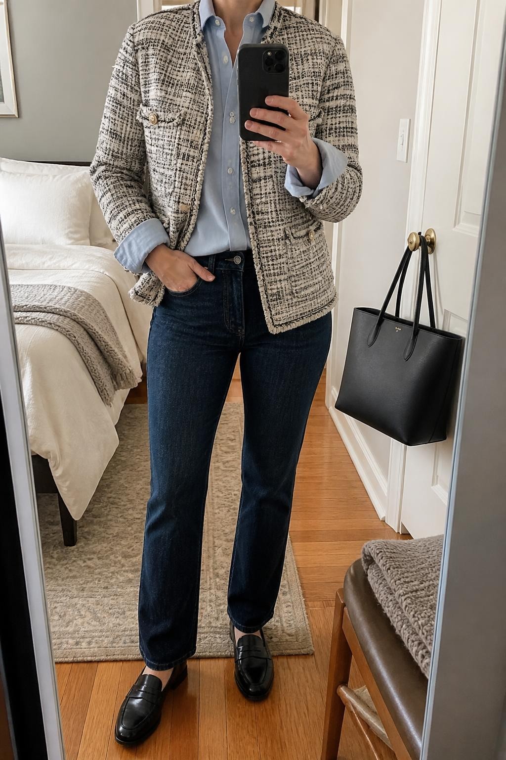 mirror selfie of a woman wearing a cream and black textured tweed jacket over a pale blue cotton button-down shirt with the cuffs visible, clean dark-wash straight-leg jeans without distressing, black leather penny loafers, and a black structured work tote, clearly showing the exact clothing items, colors, fabrics, shoes, and bag listed in the Outfit Pieces section, holding a phone in front of a mirror, cropped at NECK so the face does not show, relaxed business casual selfie pose in a cozy bedroom mirror setup, natural window light, with a handbag hanging on a door handle and a folded blanket on a chair, realistic tweed, cotton, denim, and leather textures, latest iPhone photo quality