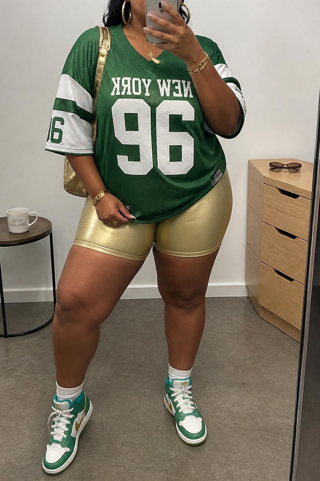 Mirror selfie of a plus size woman wearing an oversized green and white vintage-style football jersey, fitted gold metallic biker shorts, green high-top sneakers, a small gold nylon backpack, and chunky gold hoop earrings, clearly showing the exact clothing items, jersey colors, metallic shorts, footwear, and accessories listed, holding a phone in front of a mirror, cropped at the neck so her face does not show, relaxed sporty selfie pose, in a minimal dressing room with soft indoor lighting, a coffee mug on a side table and sunglasses placed on a dresser, realistic sporty festival outfit, latest iPhone photo quality.