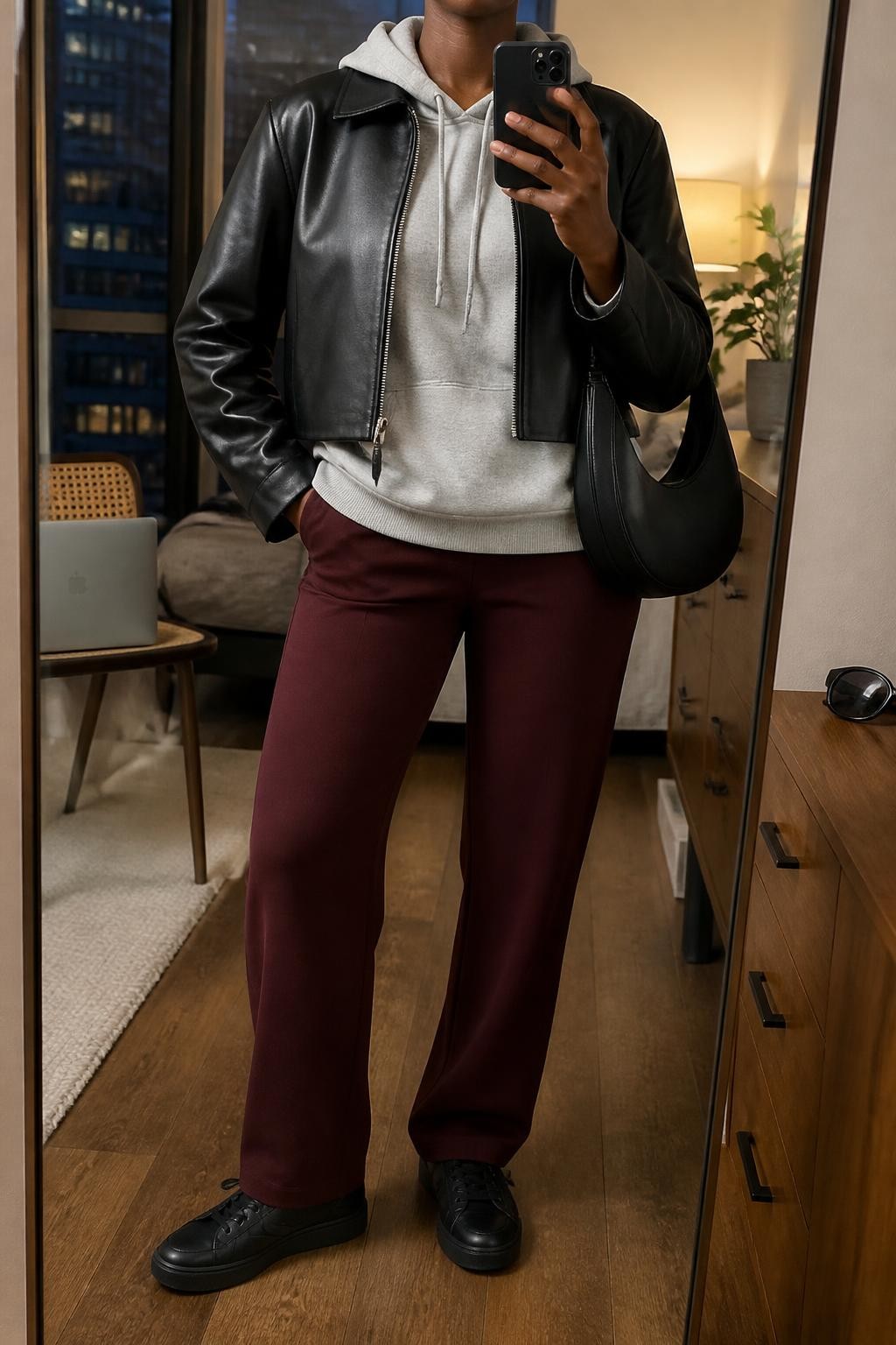 mirror selfie of a Black woman wearing an open black cropped faux-leather jacket over a heather gray cotton hoodie with the hoodie hem extending slightly below the jacket, burgundy straight-leg ponte trousers, black leather sneakers, and a black crescent shoulder bag, clearly showing the exact clothing items, colors, fabrics, sneakers, and bag listed in the Outfit Pieces section, holding a phone in front of a mirror, cropped at NECK so the face does not show, relaxed confident selfie pose in a stylish city apartment, warm moody lighting, with a laptop resting on a chair and sunglasses placed on a dresser, realistic faux-leather, cotton, ponte, and leather textures, latest iPhone photo quality