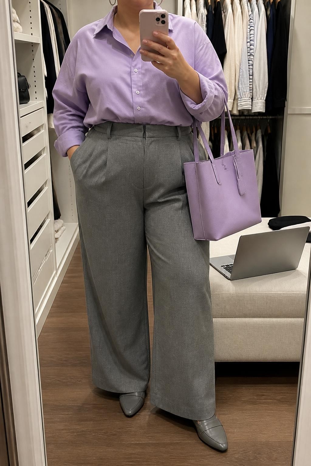 Mirror selfie of a plus size woman wearing an oversized lavender cotton button-down shirt tucked loosely into medium-grey high-waisted wide-leg tailored trousers, grey pointed loafers, a structured lavender work tote, and silver hoop earrings, clearly showing the exact clothing items, lavender cotton, grey tailoring, footwear, and accessories listed, holding a phone in front of a mirror, cropped at the neck so her face does not appear, relaxed professional selfie stance, in a walk-in closet mirror with soft indoor lighting, a laptop resting on a bench and an open closet with clothes visible, realistic office outfit, latest iPhone photo quality.