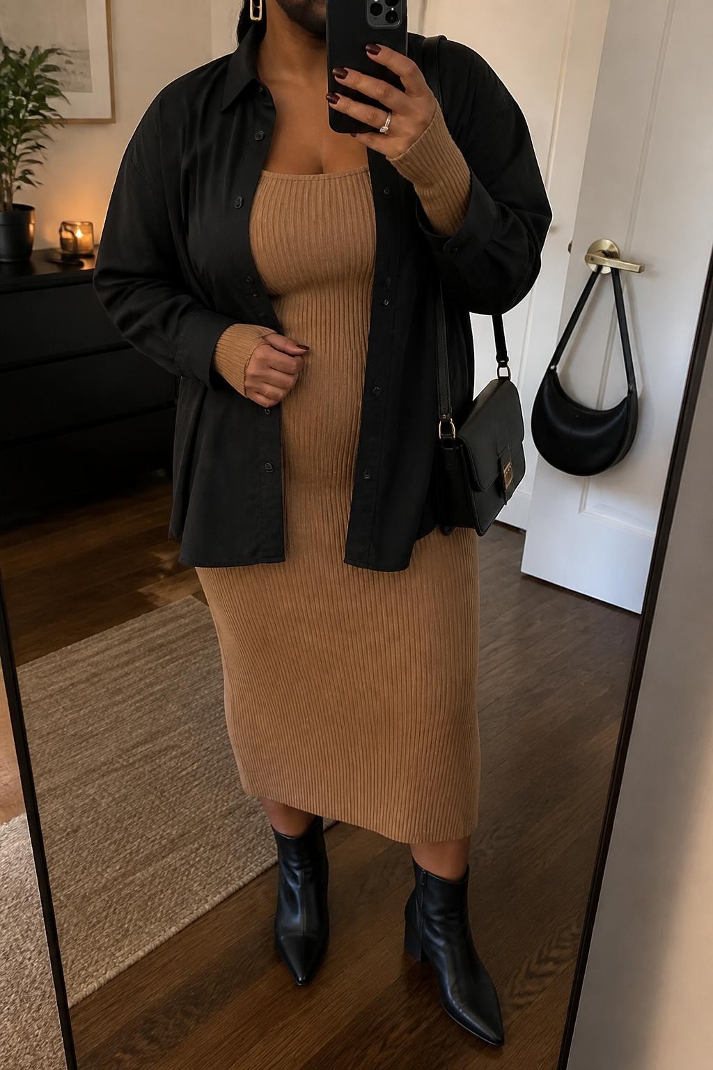 Mirror selfie of a plus size woman wearing an oversized black cotton button-down shirt worn open over a fitted camel ribbed knit midi dress with long sleeves and a square neckline, black pointed ankle boots with low block heels, a structured black shoulder bag, and gold rectangular earrings, clearly showing the exact clothing layers, black cotton, camel knit dress, footwear, and accessories listed, holding a phone in front of a mirror, cropped at the neck so the face does not show, relaxed polished selfie pose, in a stylish city apartment with warm moody lighting, a small plant on a dresser and a handbag hanging on a door handle, realistic autumn outfit, latest iPhone photo quality.