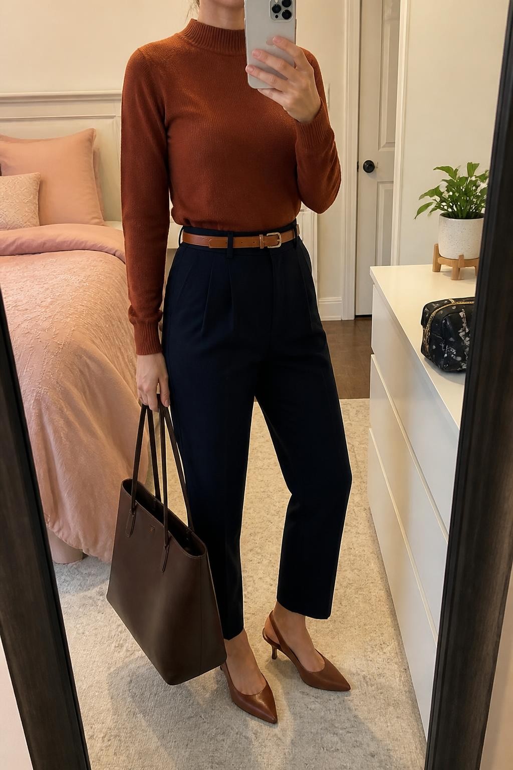 mirror selfie of a woman wearing a rust fine-knit mock-neck sweater tucked into navy high-waisted tapered trousers, a slim cognac leather belt, cognac pointed-toe slingback heels, and a dark brown leather work tote, clearly showing the exact clothing items, colors, fabrics, belt, shoes, and bag listed in the Outfit Pieces section, holding a phone in front of a mirror, cropped at NECK so the face does not show, relaxed professional selfie pose in a feminine-themed bedroom, soft indoor lighting, with a small plant on a dresser and a cosmetic bag on the counter, realistic knit, trouser, and leather textures, latest iPhone photo quality