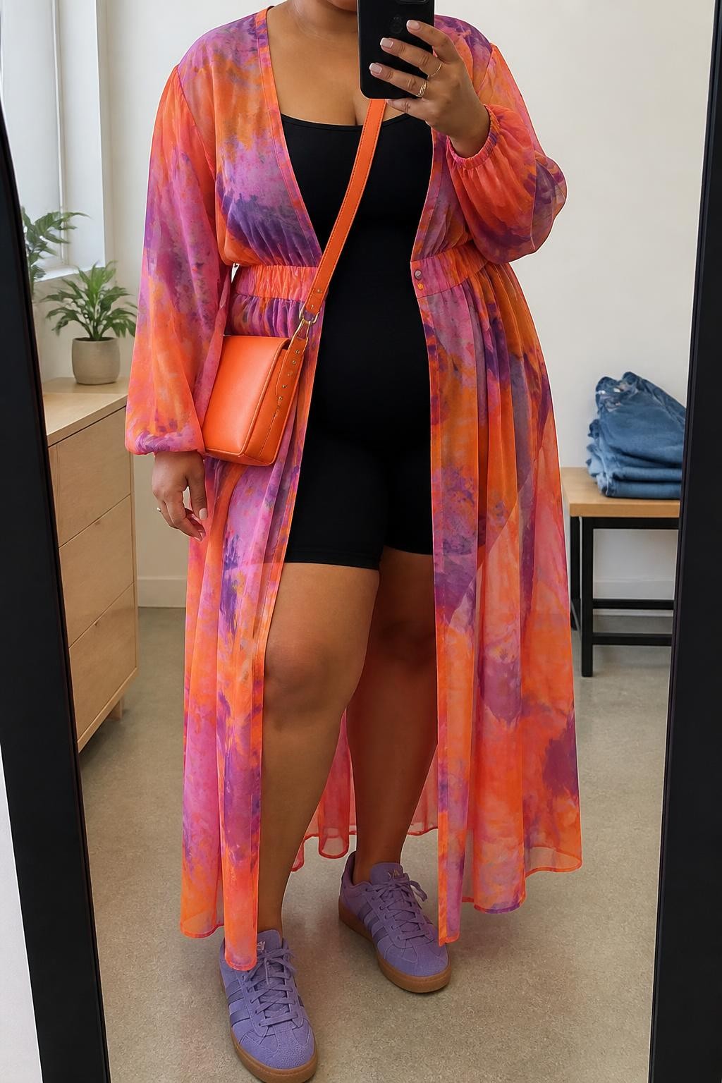 A realistic fashion mirror selfie of a plus-size adult woman wearing a colorful festival outfit: a long-sleeve maxi dress in an orange, pink, and purple abstract print, layered over a fully opaque fitted black bodysuit and black biker shorts. The dress has a lightweight, semi-transparent fashion fabric while providing full coverage, with the black layers clearly functioning as the outfit underneath. She wears purple lace-up sneakers and a small orange crossbody shoulder bag. She is holding a smartphone in front of a full-length mirror, with the composition cropped just below the chin so her face is not visible. Relaxed festival-inspired selfie pose in a minimal dressing room, natural window light, small plant on a dresser, and folded denim jeans on a bench. Authentic contemporary street-style photography, realistic fabric textures, vibrant colors, natural proportions, and high-end smartphone photo quality.