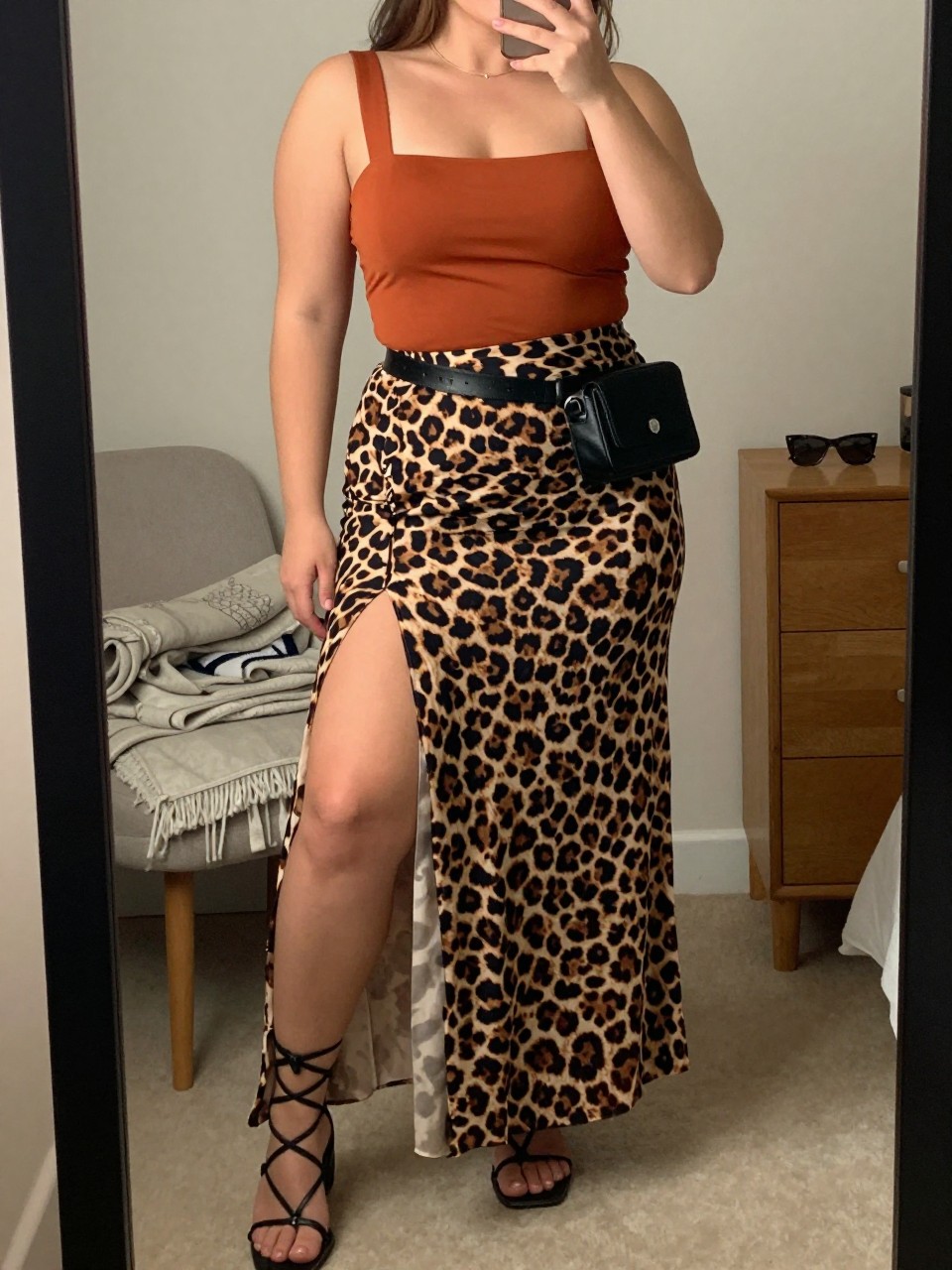 Mirror selfie of a plus size woman wearing a leopard print high-waisted chiffon maxi skirt with a thigh-high side slit and opaque lining, a fitted rust orange bandeau-style top with wide supportive straps, black flat gladiator sandals, and a small black leather belt bag, clearly showing the exact clothing items, leopard print, rust top, side slit, footwear, and accessories listed, holding a phone in front of a mirror, cropped at the neck so the face does not appear, relaxed bold selfie pose, in a cozy bedroom mirror setup with soft indoor lighting, a folded blanket on a chair and sunglasses placed on a dresser, realistic warm-weather Coachella outfit, latest iPhone photo quality.