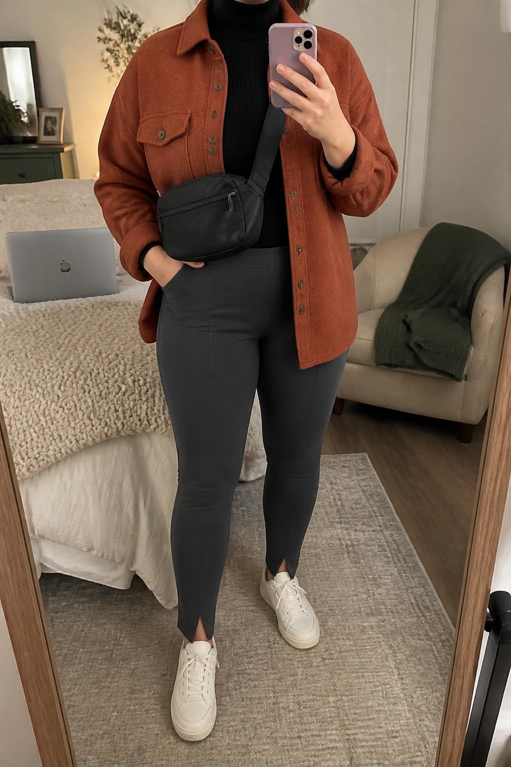 mirror selfie of a midsize woman wearing an open rust brushed wool-blend shacket over a black ribbed mock-neck top, charcoal split-hem ponte leggings, cream leather sneakers, and a black nylon crossbody bag adjusted to sit close to the waist, clearly showing the exact clothing items, colors, fabrics, sneakers, and bag listed in the Outfit Pieces section, holding a phone in front of a mirror, cropped at NECK so the face does not show, relaxed casual selfie pose in a cozy bedroom mirror setup, soft indoor lighting, with a folded blanket on a chair and a laptop on the bed, realistic wool, ribbed knit, ponte, leather, and nylon textures, slightly imperfect casual photo, latest iPhone photo quality