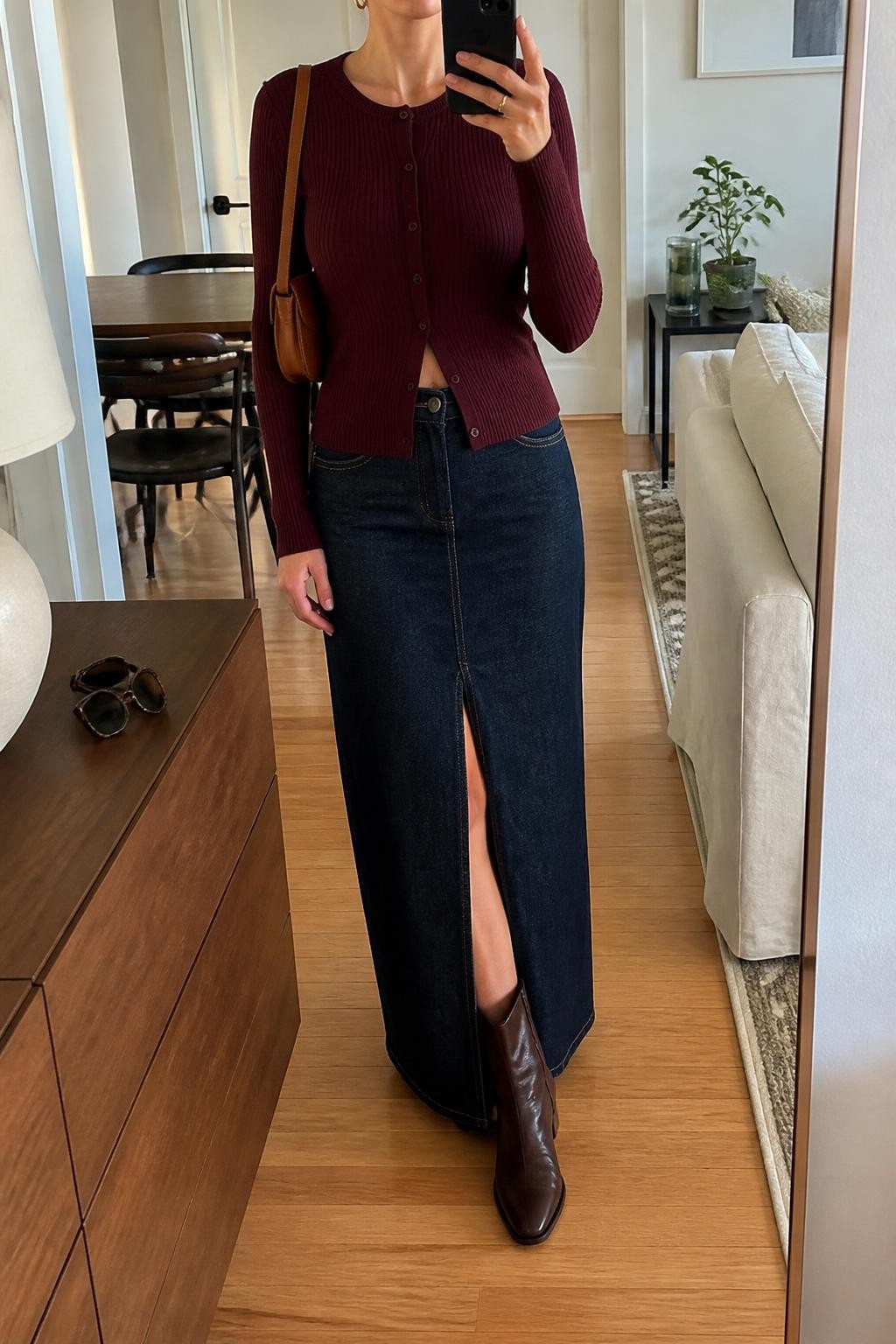 Mirror selfie of a woman wearing a dark indigo denim maxi skirt with a front slit, fitted burgundy ribbed button-front cardigan, chocolate-brown leather ankle boots, structured tan shoulder bag, and small gold hoop earrings, clearly showing the exact clothing items, colors, fabrics, and accessories listed, holding a phone in front of a stylish city apartment mirror, cropped at the neck so the face does not show, relaxed casual selfie pose, natural window light, sunglasses placed on a dresser and a small plant on a side table, natural and uncluttered, latest iPhone photo quality.