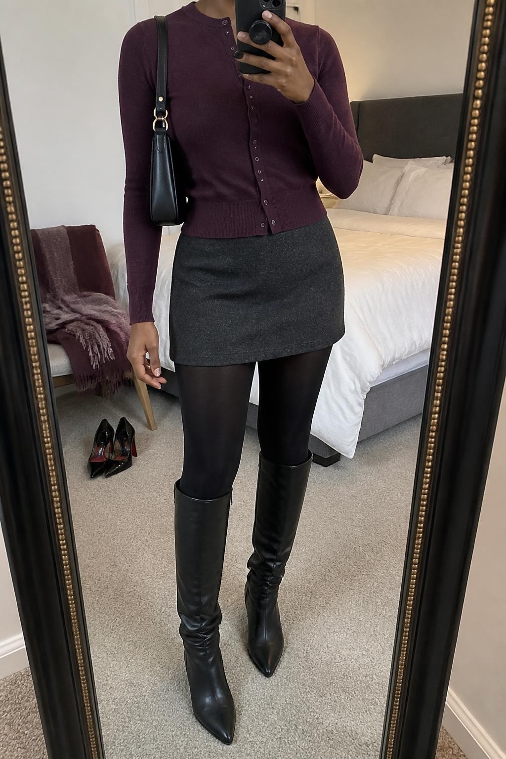 Mirror selfie of a Black woman wearing a fitted plum-purple cardigan, a charcoal-grey wool mini skirt, opaque black tights, black leather knee-high boots, and a structured black shoulder bag, clearly showing the exact clothing items, colors, fabrics, and accessories listed, holding a phone in front of a cozy bedroom mirror setup, cropped at the neck so the face does not show, relaxed casual selfie pose, soft indoor lighting, a folded blanket on a chair and a pair of heels on the floor nearby, natural lived-in atmosphere, latest iPhone photo quality.