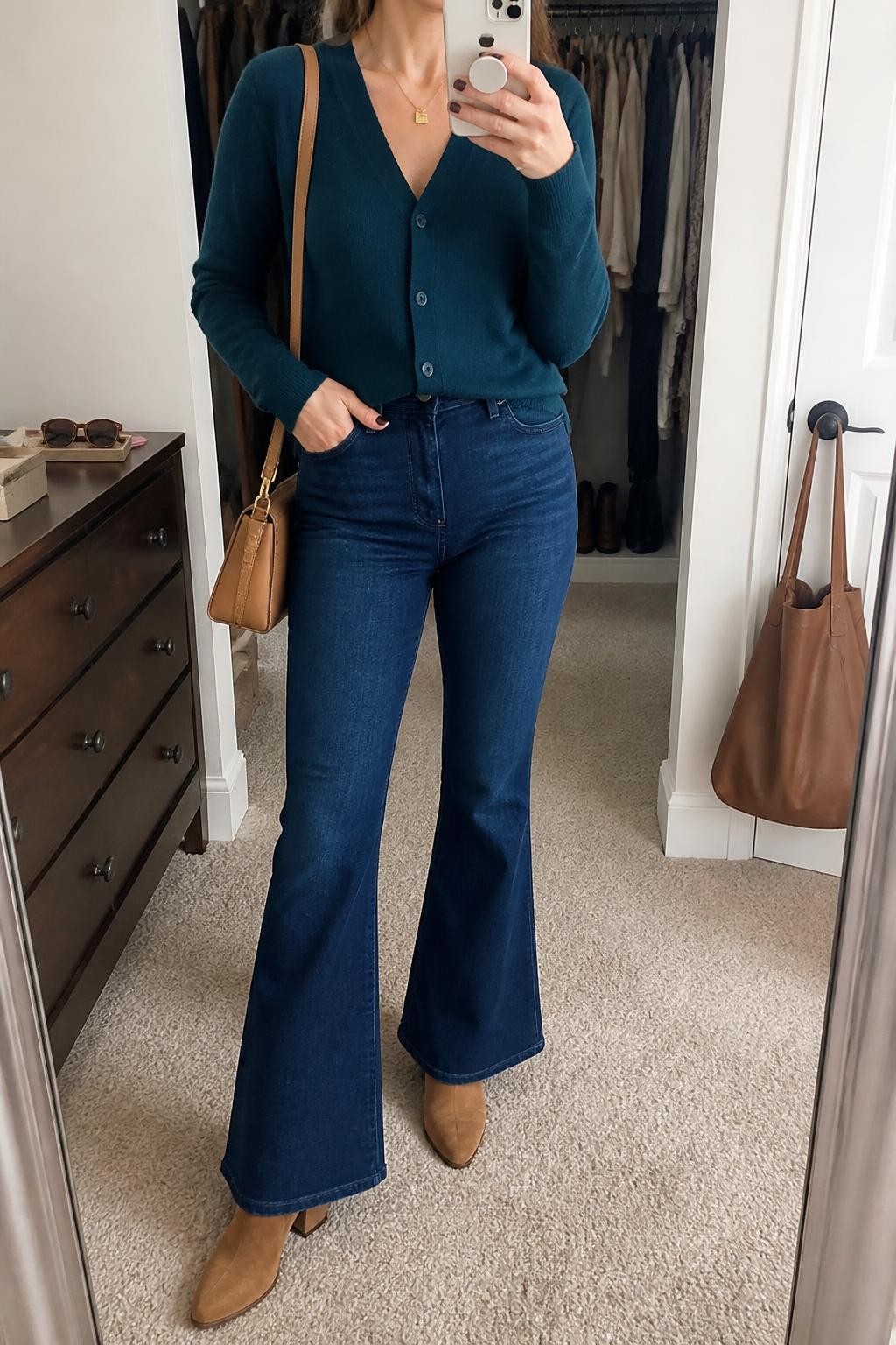 Mirror selfie of a woman wearing a deep teal V-neck cardigan, dark blue high-waisted flared jeans, brown suede block-heel ankle boots, a structured camel shoulder bag, and a delicate gold pendant necklace, clearly showing the exact clothing items, colors, fabrics, and accessories listed, holding a phone in front of a walk-in closet mirror, cropped at the neck so the face does not show, relaxed casual selfie pose, soft indoor lighting, sunglasses placed on a dresser and a handbag hanging on a door handle, natural everyday setting, latest iPhone photo quality.