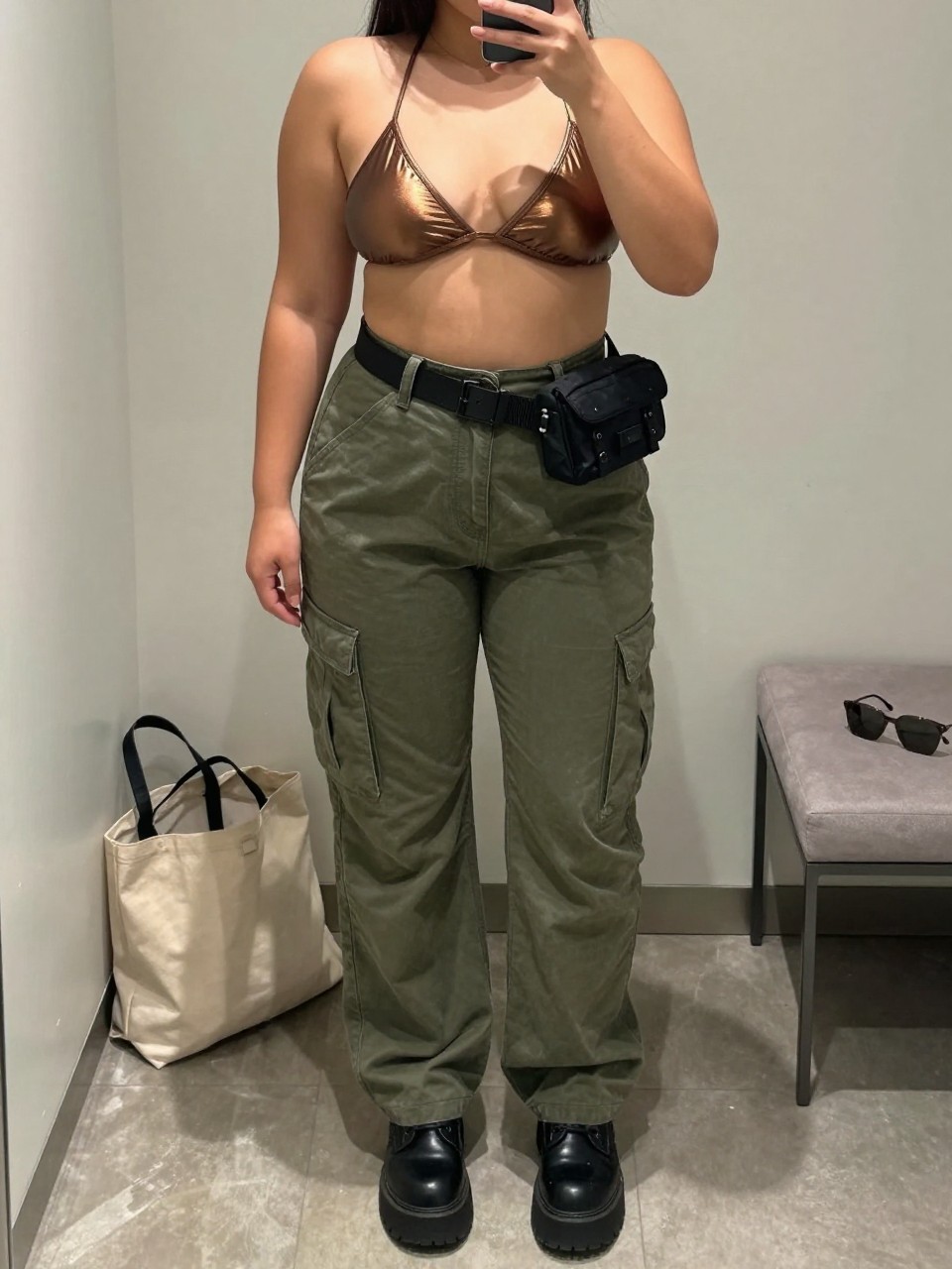 Mirror selfie of a plus size woman wearing olive green high-waisted cargo pants with relaxed straight legs and flat utility pockets, a metallic bronze bikini-style top with wide supportive straps and full coverage, black platform combat boots, and a compact black nylon belt bag, clearly showing the exact clothing items, olive cargo fabric, bronze top, black footwear, and accessories listed, holding a phone in front of a mirror, cropped at the neck so the face does not appear, relaxed edgy selfie pose, in a minimal dressing room with soft indoor lighting, a tote bag resting on the floor and sunglasses placed on a bench, realistic Coachella styling, latest iPhone photo quality.