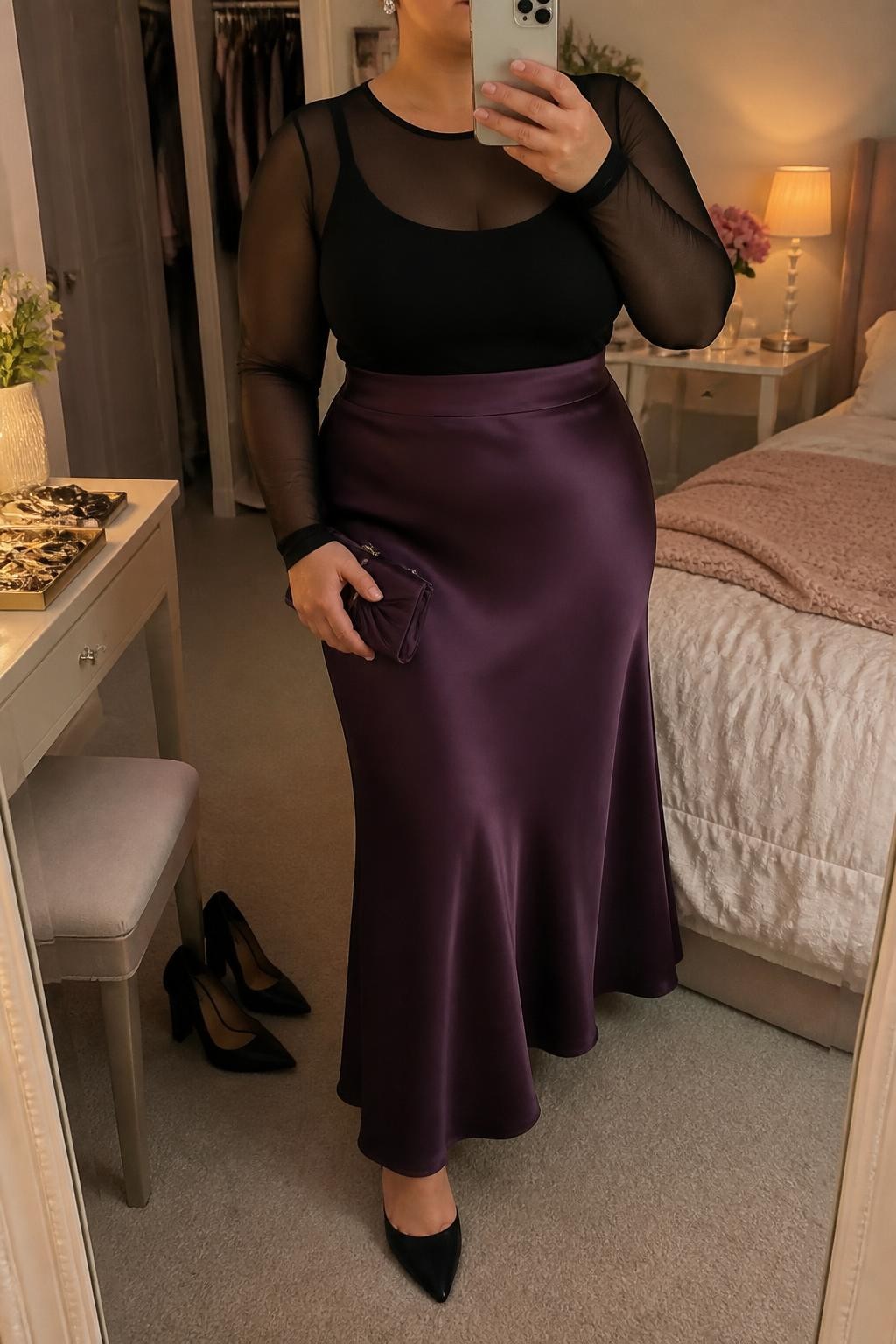 Mirror selfie of a plus size woman wearing a plum purple satin high-waisted maxi skirt with a fluid bias-cut shape, a fitted black mesh long-sleeve top layered over a full-coverage black singlet, black pointed block heels, a small plum satin clutch, and silver crystal stud earrings, clearly showing the exact clothing layers, plum satin, black mesh, colors, footwear, and accessories listed, holding a phone in front of a mirror, cropped at the neck so the face is hidden, relaxed glamorous selfie pose, in a feminine-themed bedroom with warm moody lighting, a pair of heels on the floor nearby and a jewelry tray on the vanity, realistic evening outfit, latest iPhone photo quality.