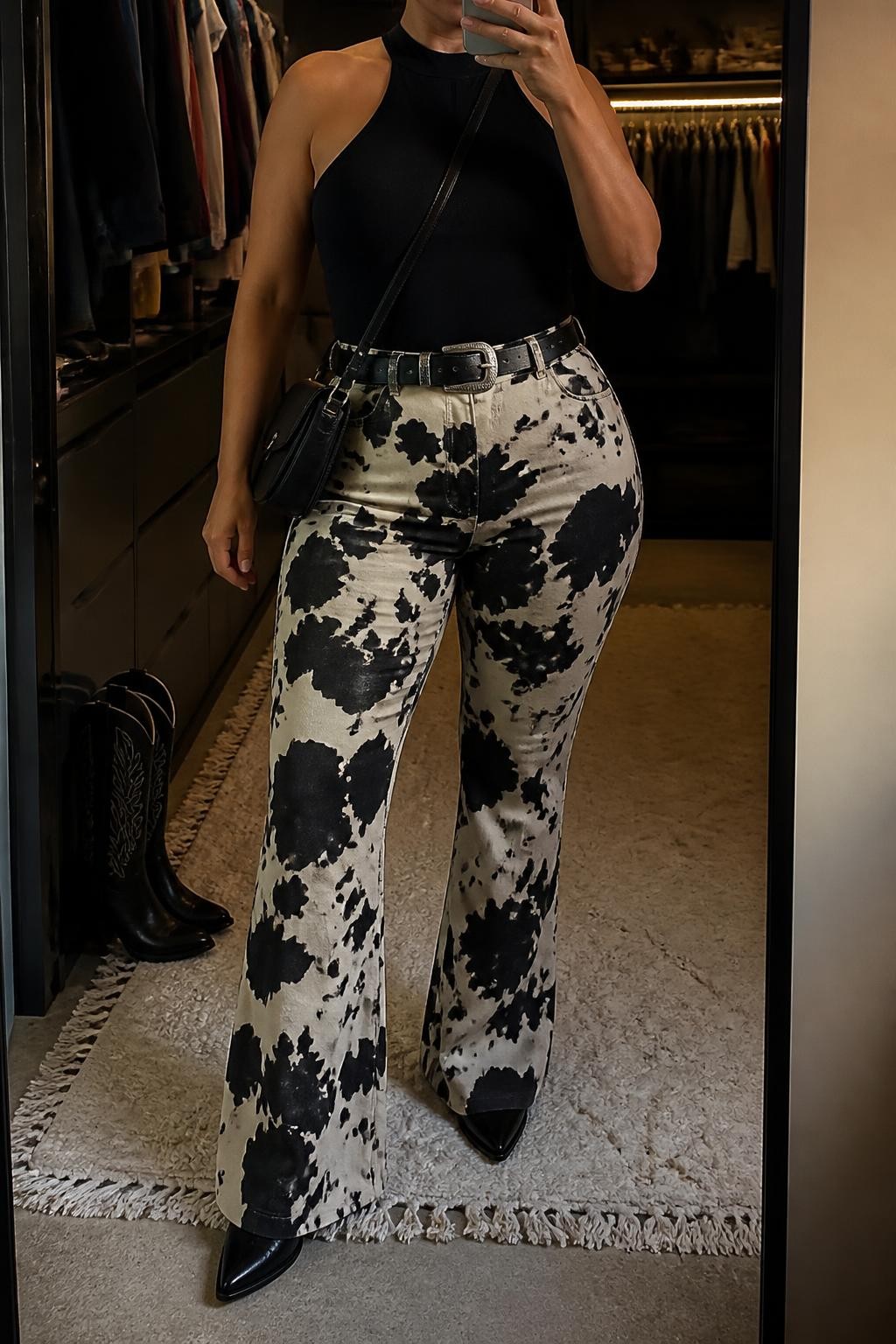 Mirror selfie of a plus size woman wearing black and cream cow-print high-waisted flared pants in stretch fabric, a fitted black halter-neck top with a wide supportive neck band, black pointed ankle cowboy boots, a black leather Western belt with a silver buckle, and a compact black crossbody bag, clearly showing the exact clothing items, cow print, black fitted top, footwear, belt, and accessories listed, holding a phone in front of a mirror, cropped at the neck so the face is hidden, relaxed confident selfie pose, in a walk-in closet mirror with warm moody lighting, an open closet with clothes visible and a pair of boots on the floor nearby, realistic Coachella outfit, latest iPhone photo quality.