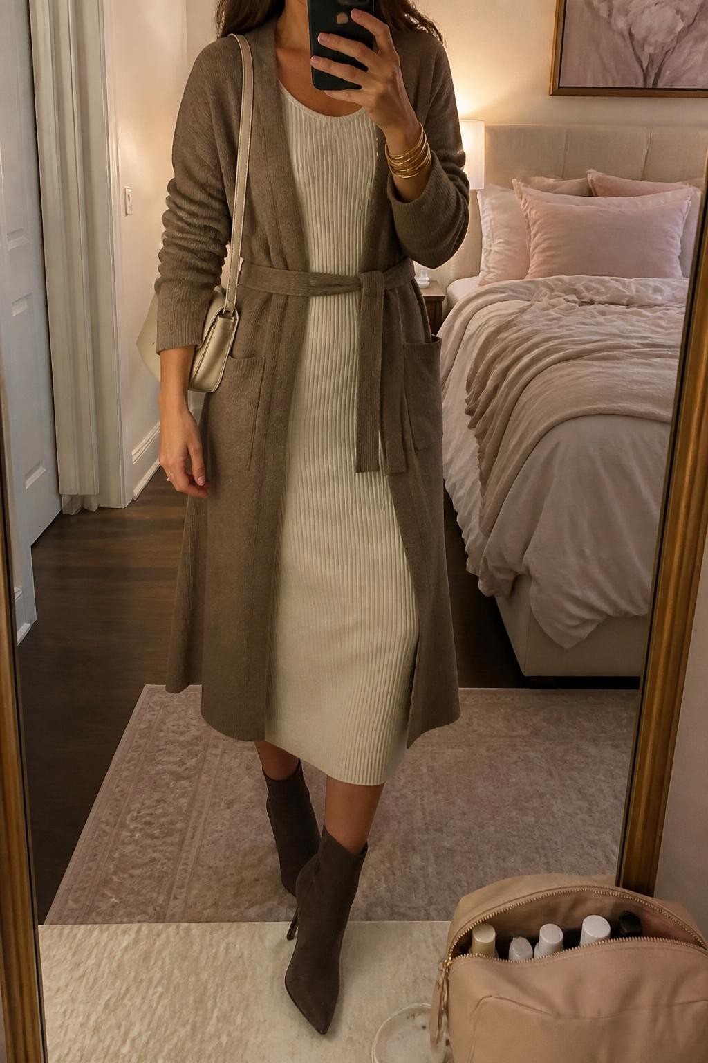 Mirror selfie of a woman wearing a long mocha-brown belted cardigan over a fitted ivory ribbed-knit midi sweater dress, chocolate-brown suede ankle boots, a cream leather shoulder bag, and thin gold bangles, clearly showing the exact clothing items, colors, fabrics, and accessories listed, holding a phone in front of a feminine-themed bedroom mirror, cropped at the neck so the face does not show, relaxed casual selfie pose, warm moody lighting, soft pillows on an unmade bed and a cosmetic bag on the counter, cozy and uncluttered, latest iPhone photo quality.