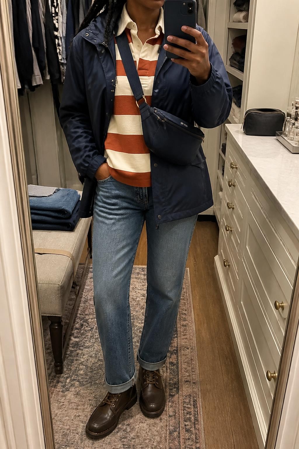 mirror selfie of a Black woman wearing an open navy water-resistant hooded rain jacket over a cream and rust striped cotton rugby shirt, medium-wash straight-leg jeans, dark brown weather-resistant lace-up boots, and a navy nylon crossbody bag, clearly showing the exact clothing items, colors, striped pattern, fabrics, boots, and bag listed in the Outfit Pieces section, holding a phone in front of a mirror, cropped at NECK so the face does not show, relaxed casual selfie pose in a walk-in closet mirror, soft indoor lighting, with folded denim jeans on a bench and a cosmetic bag on the vanity, realistic water-resistant nylon, cotton, denim, and leather textures, latest iPhone photo quality