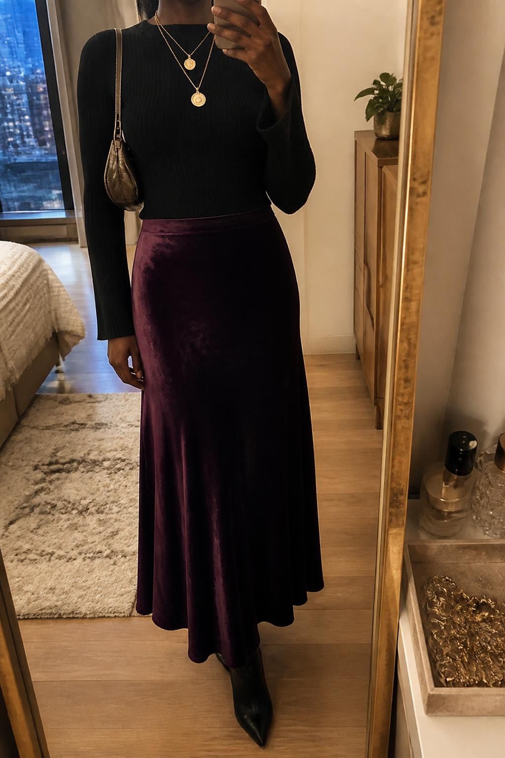 Mirror selfie of a Black woman wearing a deep plum velvet maxi skirt, a fitted black knit top with soft bell sleeves, black suede ankle boots, a metallic bronze mini shoulder bag, and layered gold pendant necklaces, clearly showing the exact clothing items, colors, fabrics, and accessories listed, holding a phone in front of a stylish city apartment mirror, cropped at the neck so the face does not show, relaxed casual selfie pose, warm moody lighting, a jewelry tray on the vanity and a small plant on a dresser, elegant natural setting, latest iPhone photo quality.