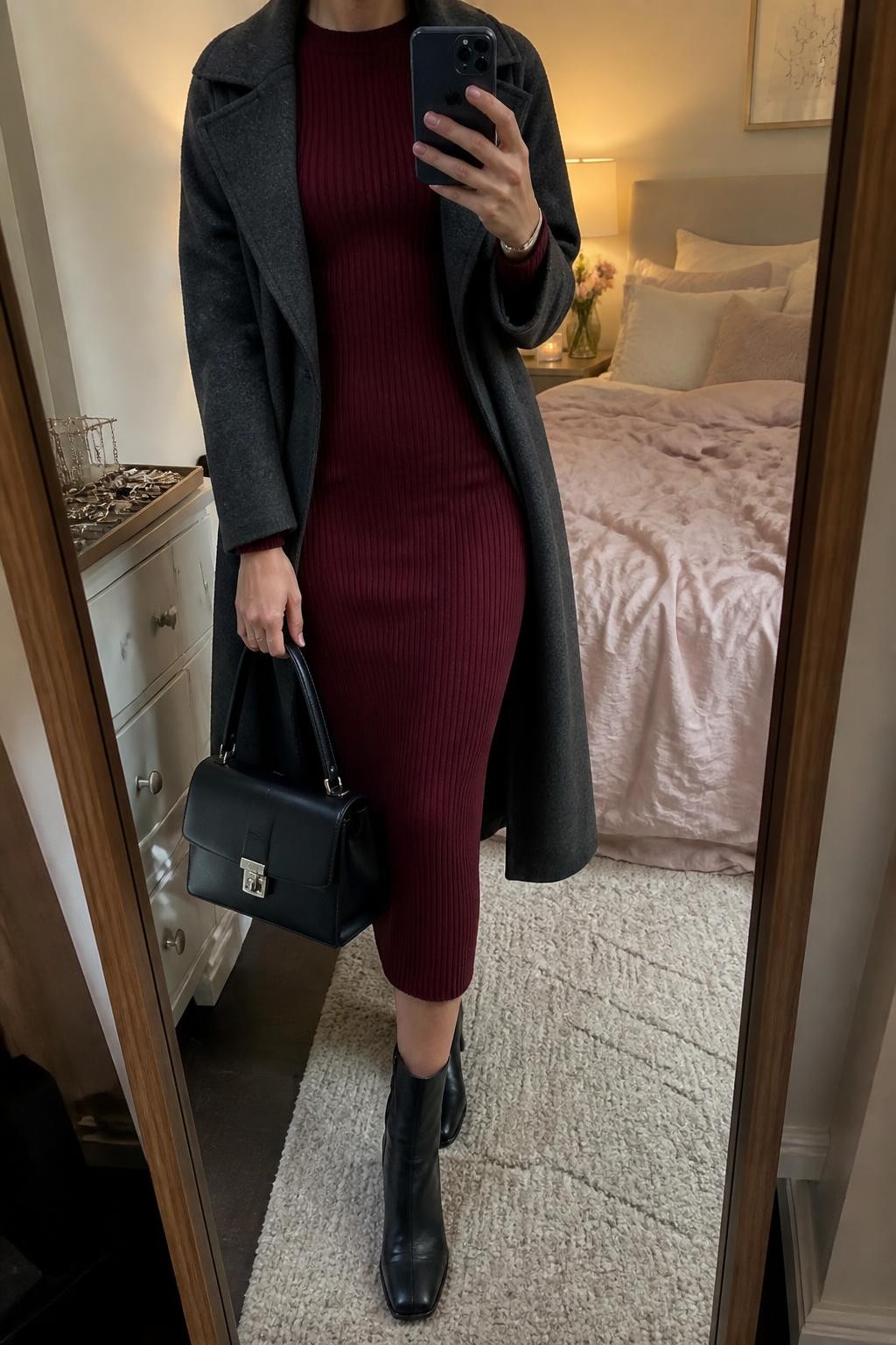mirror selfie of a woman wearing an open charcoal gray wool-blend trench coat over a burgundy ribbed long-sleeve midi dress, black leather block-heel ankle boots, a black structured top-handle bag, and a slim silver cuff bracelet, clearly showing the exact clothing items, colors, fabrics, boots, bag, and bracelet listed in the Outfit Pieces section, holding a phone in front of a mirror, cropped at NECK so the face does not show, relaxed elegant selfie pose in a feminine-themed bedroom, warm moody lighting, with soft pillows on an unmade bed and a jewelry tray on a dresser, realistic wool, ribbed knit, leather, and silver textures, latest iPhone photo quality