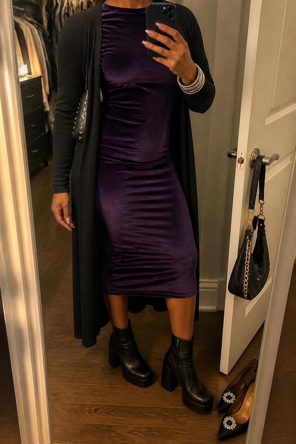 mirror selfie of a Black woman wearing a deep purple velvet long-sleeve midi dress, an open black duster cardigan, black leather platform ankle boots, a black crescent shoulder bag with small silver star studs, and layered silver bangles, clearly showing the exact clothing items, colors, fabrics, boots, bag, studs, and bracelets listed in the Outfit Pieces section, holding a phone in front of a mirror, cropped at NECK so the face does not show, relaxed confident selfie pose in a walk-in closet mirror, warm moody lighting, with a pair of heels on the floor nearby and a handbag hanging on a door handle, realistic velvet, knit, leather, and silver textures, latest iPhone photo quality