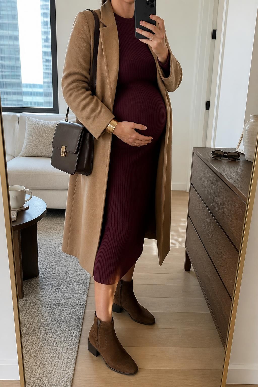 mirror selfie of a pregnant woman wearing a burgundy ribbed maternity midi dress shaped comfortably around her visible baby bump, an open camel wool-blend coat, chocolate suede low block-heel ankle boots, a dark brown structured shoulder bag, and a brushed gold cuff bracelet, clearly showing the exact clothing items, colors, fabrics, boots, bag, and bracelet listed in the Outfit Pieces section, holding a phone in front of a full-length mirror, cropped at NECK so the face does not show, relaxed natural maternity selfie pose in a stylish city apartment, natural window light, with a coffee mug on a side table and sunglasses placed on a dresser, realistic ribbed knit, wool, suede, and leather textures, natural pregnancy proportions, latest iPhone photo quality