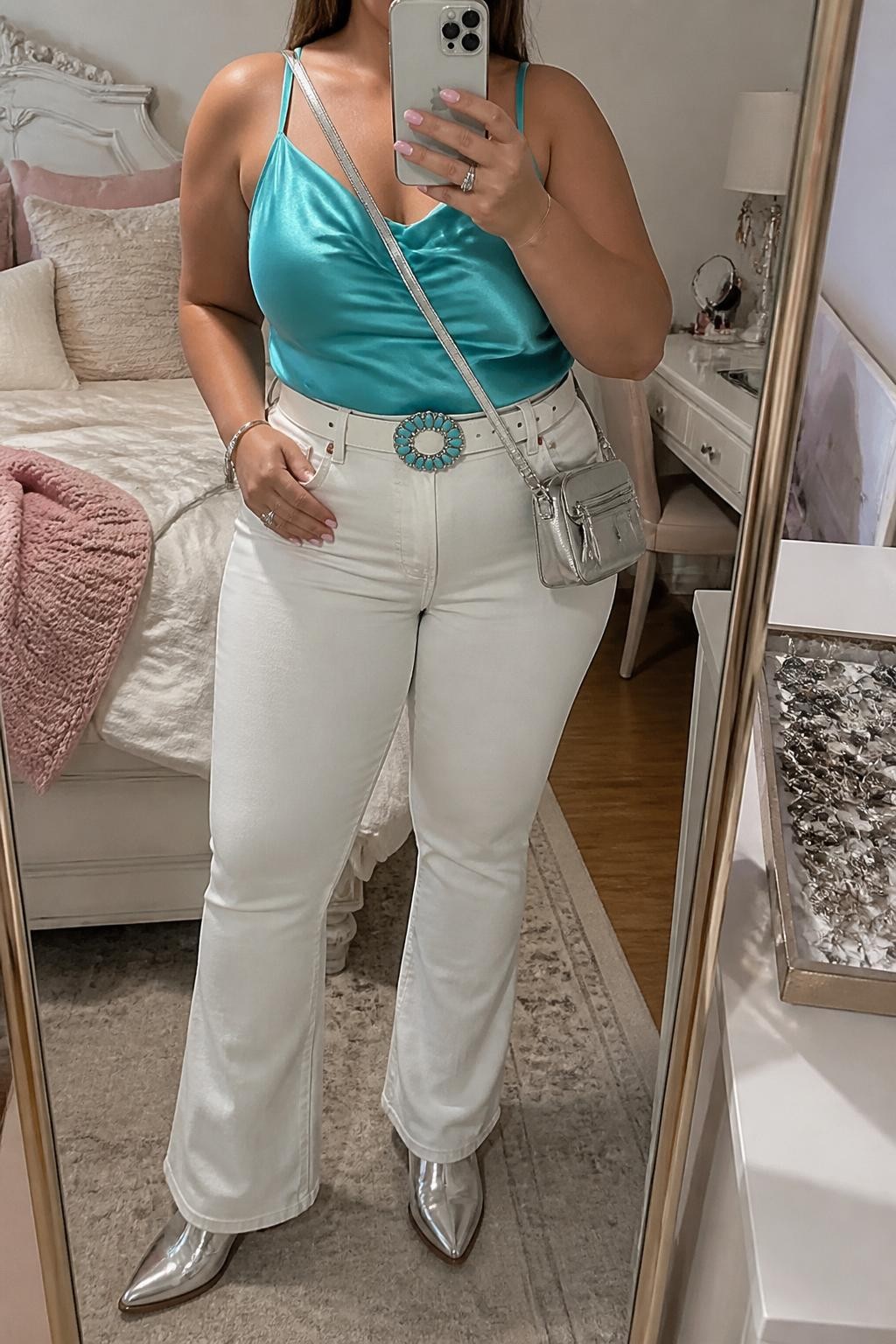 Mirror selfie of a plus size woman wearing a turquoise satin camisole with a softly draped neckline and wide supportive straps, high-waisted white flared jeans, metallic silver pointed cowboy boots, a white Western belt with a turquoise buckle, and a small silver leather crossbody bag, clearly showing the exact clothing items, turquoise satin, white denim, metallic footwear, belt, and accessories listed, holding a phone in front of a mirror, cropped at the neck so her face does not show, relaxed polished selfie stance, in a feminine-themed bedroom with soft indoor lighting, a jewelry tray on the vanity and soft pillows on an unmade bed, realistic fresh country concert outfit, latest iPhone photo quality.