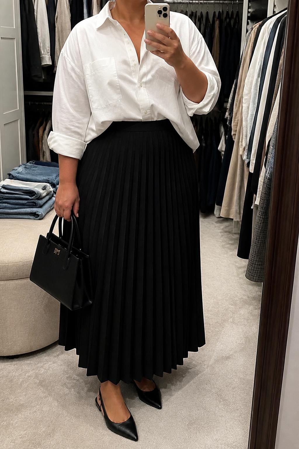 Mirror selfie of a plus size woman wearing a black high-waisted pleated maxi skirt, an oversized white cotton button-down shirt tucked loosely at the front, black pointed slingback flats, a structured black top-handle bag, and small silver hoop earrings, clearly showing the exact clothing items, black pleats, white cotton, colors, footwear, and accessories listed, holding a phone in front of a mirror, cropped at the neck so her face does not appear, relaxed polished selfie stance, in a walk-in closet mirror with soft indoor lighting, an open closet with clothes visible and folded denim jeans on a bench, realistic smart-casual styling, latest iPhone photo quality.