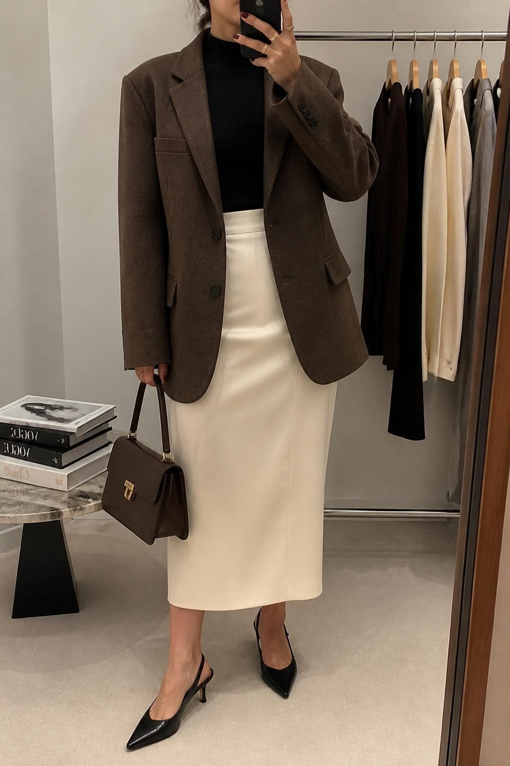 Mirror selfie of a woman wearing an oversized chocolate-brown wool blazer, a fitted black mock-neck top, a structured cream column midi skirt, black leather slingback heels, and a dark brown top-handle handbag, clearly showing the exact clothing items, colors, fabrics, and accessories listed, holding a phone in front of a minimal dressing room mirror, cropped at the neck so the face does not show, relaxed confident selfie pose, soft indoor lighting, a clothing rack with garments behind and a stack of fashion magazines on a side table, modern professional setting, latest iPhone photo quality.