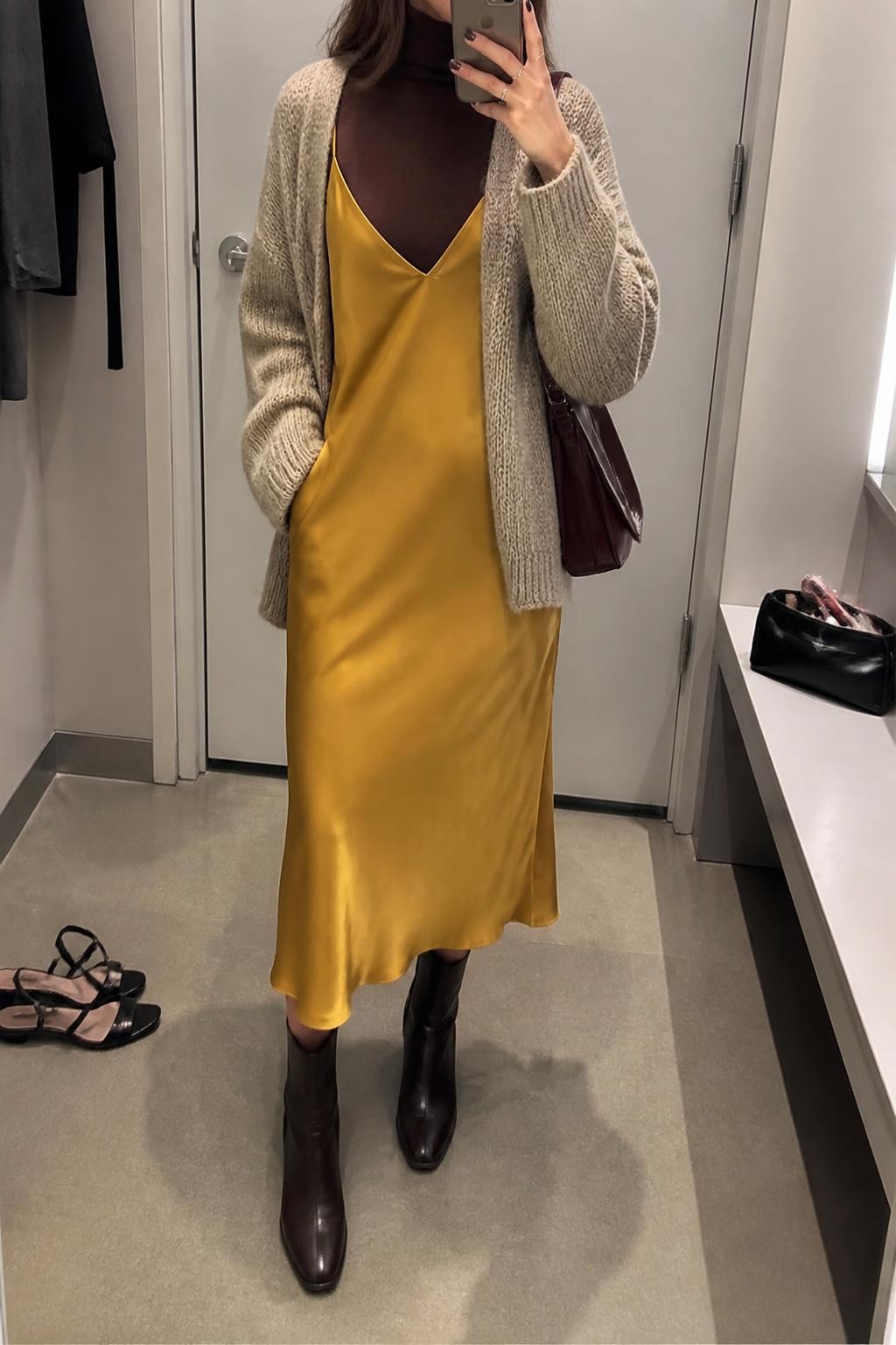 Mirror selfie of a woman wearing a mustard-yellow satin midi slip dress layered over a fitted chocolate-brown turtleneck, an oversized oatmeal chunky-knit cardigan, dark brown leather ankle boots, and a small burgundy shoulder bag, clearly showing the exact clothing items, colors, fabrics, and accessories listed, holding a phone in front of a minimal dressing room mirror, cropped at the neck so the face does not show, relaxed casual selfie pose, soft indoor lighting, a pair of heels on the floor nearby and a makeup bag on the counter, natural and unstaged, latest iPhone photo quality.