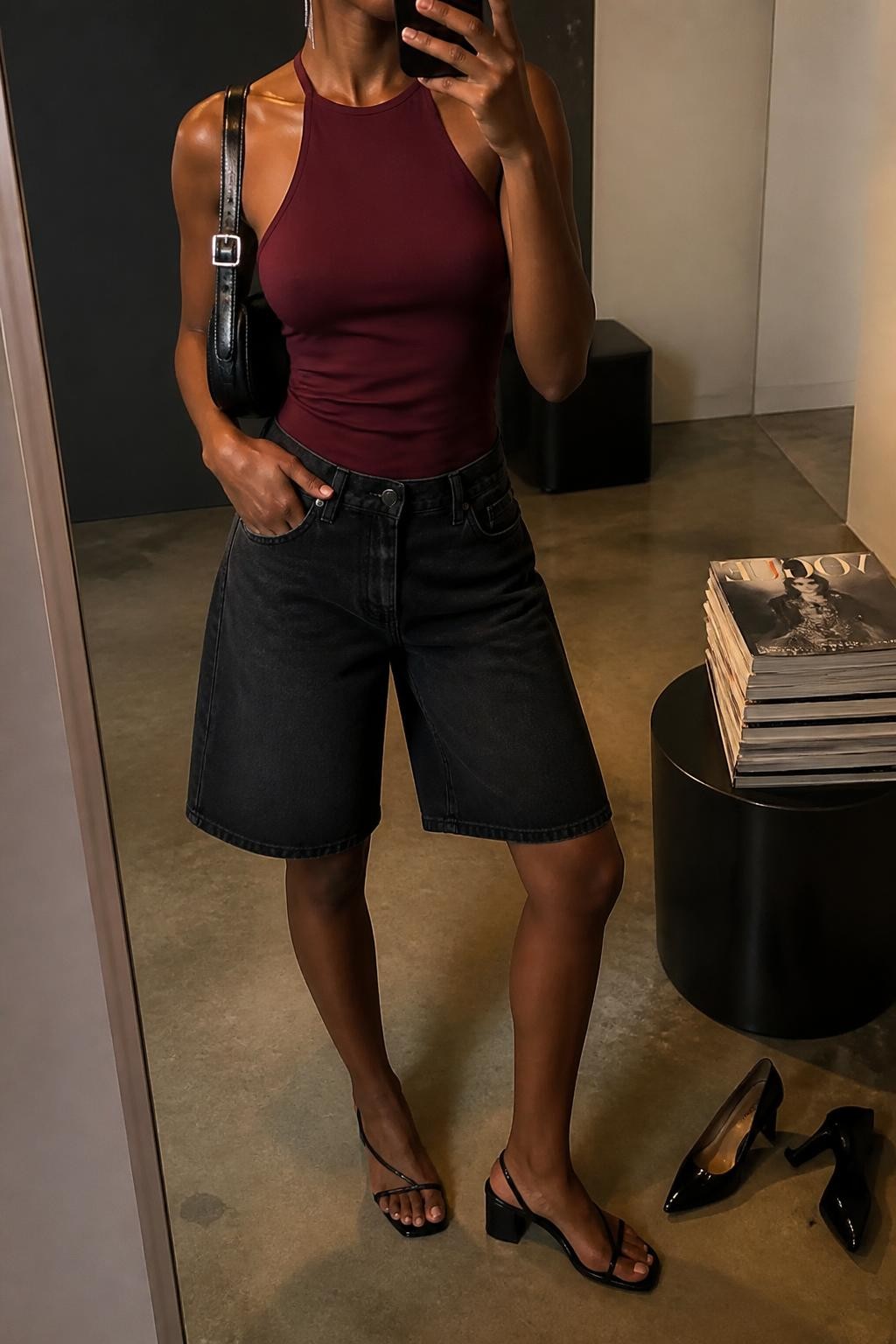 mirror selfie of a Black woman wearing relaxed straight-fit washed black denim jorts, a fitted burgundy stretch halter top, black low block-heel sandals, a black leather shoulder bag with silver hardware and slim silver drop earrings, wearing exactly the outfit listed with no extra garments or accessories, holding a phone in front of a mirror, cropped at NECK so the face does not show, relaxed casual selfie pose, in a fashion studio mirror corner, warm moody lighting, pair of heels on the floor nearby and a stack of fashion magazines on a side table, believable casual iPhone photography, latest iPhone photo quality.