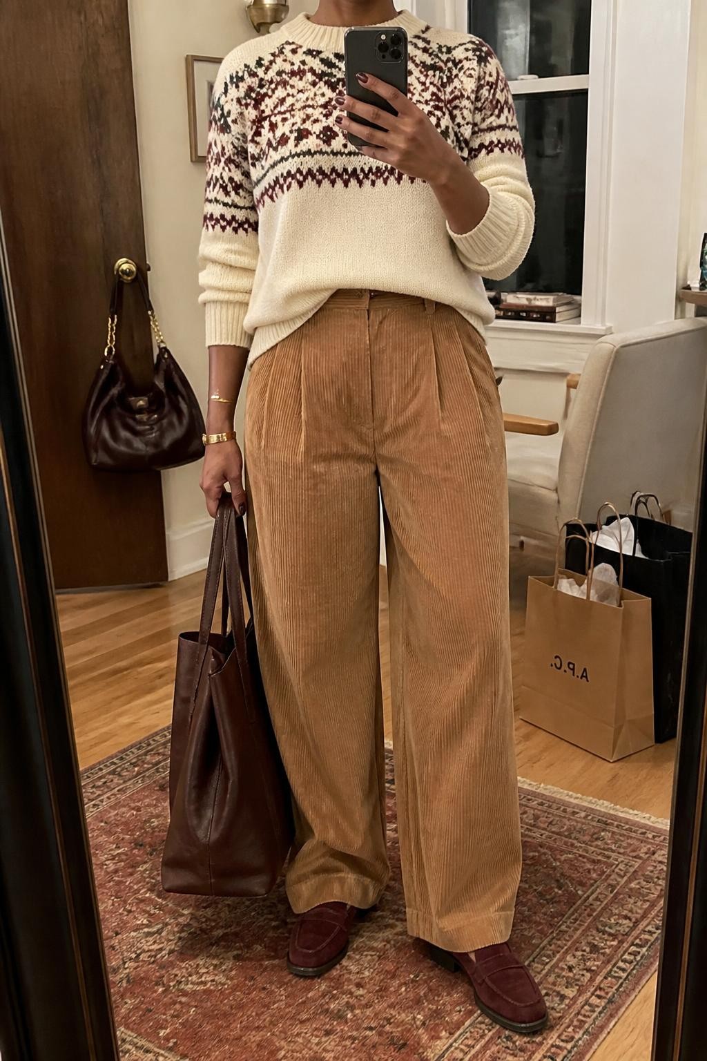 mirror selfie of a Black midsize woman wearing a cream Fair Isle sweater with a burgundy and forest green pattern, camel pleated wide-leg corduroy trousers with a small front sweater tuck, burgundy suede loafers, a dark brown leather tote bag held at her side, and a brushed gold cuff bracelet, clearly showing the exact clothing items, colors, patterns, fabrics, shoes, tote, and bracelet listed in the Outfit Pieces section, holding a phone in front of a mirror, cropped at NECK so the face does not show, relaxed casual selfie pose in a stylish city apartment, warm moody lighting, with shopping bags beside a chair and a handbag hanging on a door handle, realistic knit, corduroy, suede, and leather textures, natural fall outfit photo, latest iPhone photo quality