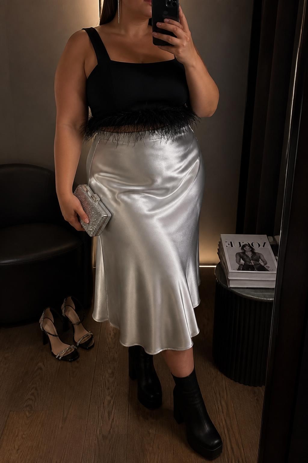 Mirror selfie of a plus size woman wearing a silver high-waisted satin midi skirt with a softly flared shape, a fitted black sleeveless square-neck top with delicate feather trim around the hem, black platform ankle boots, a small silver box clutch, and silver linear earrings, clearly showing the exact clothing items, silver satin, black feather trim, footwear, and accessories listed, holding a phone in front of a mirror, cropped at the neck so her face does not show, relaxed glamorous selfie stance, in a minimal dressing room with warm moody lighting, a pair of heels on the floor nearby and a stack of fashion magazines on a side table, realistic party outfit, latest iPhone photo quality.