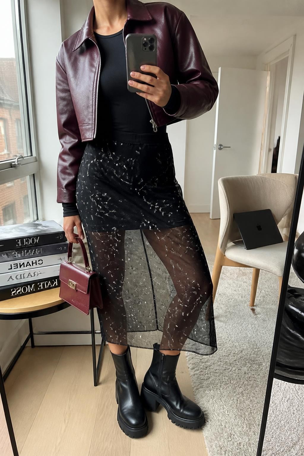 mirror selfie of a woman wearing an open oxblood cropped faux-leather jacket over a black fitted long-sleeve bodysuit tucked into a lined black mesh midi skirt with clear silver constellation embroidery, black chunky ankle boots, and an oxblood structured mini bag, clearly showing the exact clothing items, colors, fabrics, embroidery, boots, and bag listed in the Outfit Pieces section, holding a phone in front of a mirror, cropped at NECK so the face does not show, relaxed bold selfie pose in a stylish city apartment, natural window light, with a stack of fashion magazines on a side table and a laptop on a chair, realistic faux-leather, bodysuit fabric, mesh, embroidery, and leather textures, latest iPhone photo quality