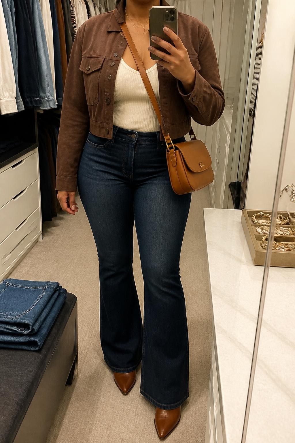 mirror selfie of a Black curvy woman wearing a chocolate brown cropped suede jacket over a cream fitted ribbed scoop-neck top, dark-wash high-rise flare jeans lightly covering cognac leather heeled ankle boots, and a tan leather crossbody bag, clearly showing the exact clothing items, colors, fabrics, boots, and bag listed in the Outfit Pieces section, holding a phone in front of a mirror, cropped at NECK so the face does not show, relaxed casual selfie pose in a walk-in closet mirror, soft indoor lighting, with folded denim jeans on a bench and a jewelry tray on the vanity, realistic suede, ribbed knit, denim, and leather textures, latest iPhone photo quality
