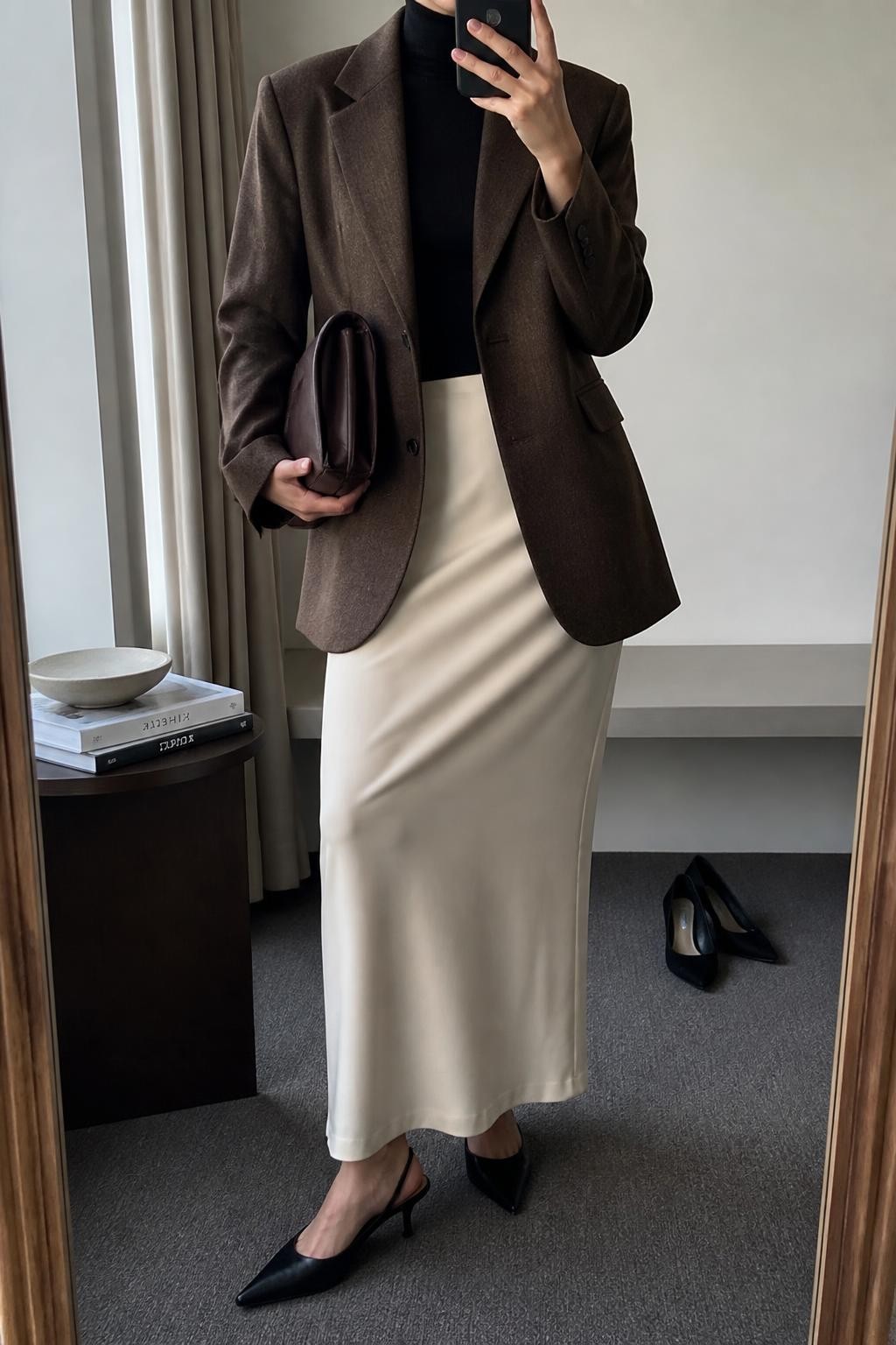 Mirror selfie of a woman wearing a cream column maxi skirt, oversized cocoa-brown wool blazer, fitted black turtleneck, black leather slingback heels, and chocolate-brown clutch bag, clearly showing the exact clothing items, colors, fabrics, and accessories listed, holding a phone in front of a minimal dressing room mirror, cropped at the neck so the face does not show, relaxed casual selfie pose, natural window light, pair of heels on the floor nearby and a stack of fashion magazines on a side table, modern natural setting, latest iPhone photo quality.