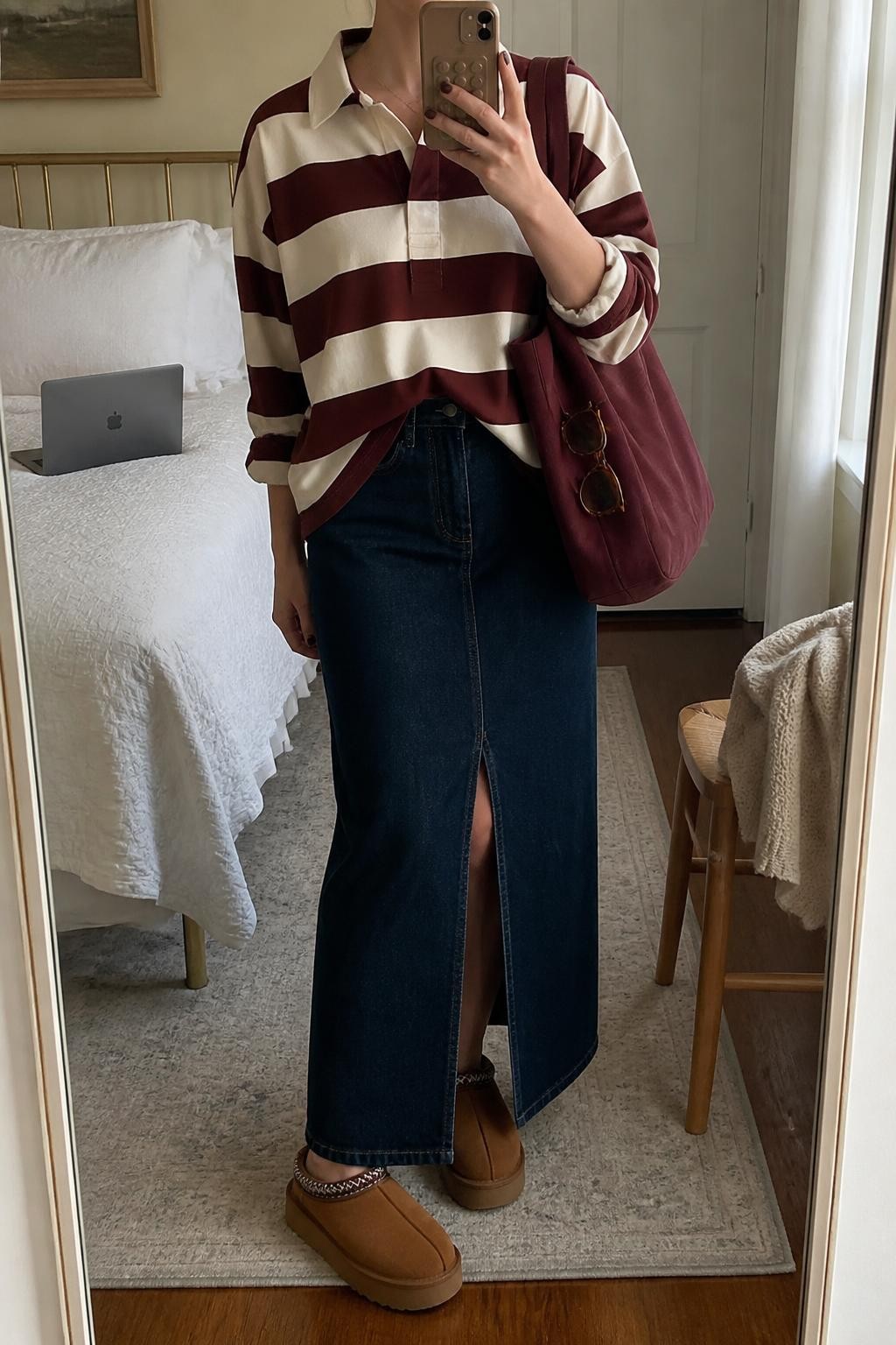 mirror selfie of a woman wearing a cream and burgundy striped cotton rugby shirt with a small front tuck into a dark-wash front-slit denim maxi skirt, chestnut Tazz platform Ugg clogs, a burgundy canvas tote bag, and tortoiseshell rectangular sunglasses hooked onto the tote, clearly showing the exact clothing items, colors, pattern, fabrics, clogs, bag, and sunglasses listed in the Outfit Pieces section, holding a phone in front of a mirror, cropped at NECK so the face does not show, relaxed casual selfie pose in a cozy bedroom mirror setup, natural window light, with a laptop on the bed and a folded blanket on a chair, realistic cotton, denim, suede, canvas, and tortoiseshell textures, latest iPhone photo quality