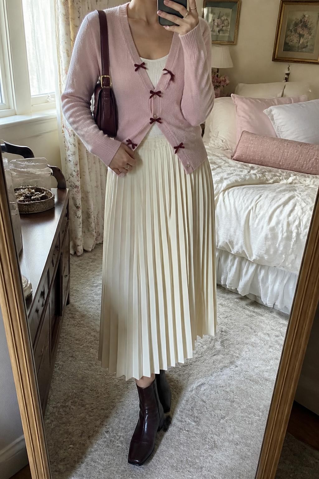 Image Prompt mirror selfie of a woman wearing a blush pink fine-knit cardigan with small burgundy velvet bows buttoned only through the middle over an ivory fitted scoop-neck knit top, a cream pleated midi skirt, chocolate brown leather ankle boots, and a burgundy structured shoulder bag, clearly showing the exact clothing items, colors, fabrics, bows, boots, and bag listed in the Outfit Pieces section, holding a phone in front of a full-length mirror, cropped at NECK so the face does not show, relaxed feminine selfie pose in a feminine-themed bedroom, natural window light, with soft pillows on an unmade bed and a jewelry tray on a dresser, realistic knit, velvet, pleated fabric, and leather textures, natural unstaged mirror photo, latest iPhone photo quality