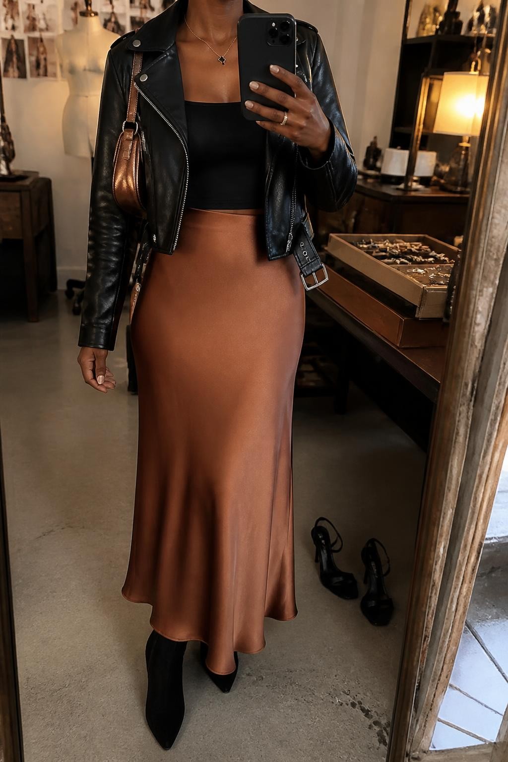 Mirror selfie of a Black woman wearing a copper satin maxi skirt, cropped black leather moto jacket, fitted black square-neck top, black suede ankle boots, and metallic bronze mini shoulder bag, clearly showing the exact clothing items, colors, fabrics, and accessories listed, holding a phone in front of a fashion studio mirror corner, cropped at the neck so the face does not show, relaxed casual selfie pose, warm moody lighting, jewelry tray on the vanity and a pair of heels on the floor nearby, natural editorial atmosphere, latest iPhone photo quality.