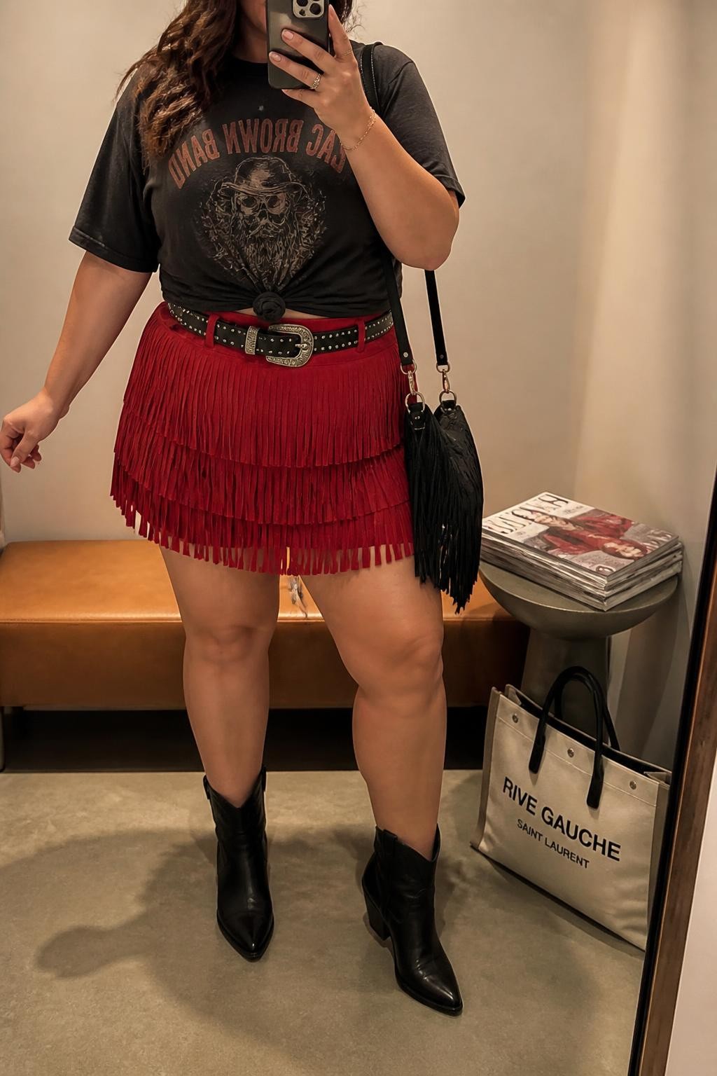 Mirror selfie of a plus size woman wearing a faded black oversized country band T-shirt tied gently at the waist, a high-waisted red suede mini skirt with layered fringe, black leather ankle cowboy boots, a black studded Western belt, and a small black fringe shoulder bag, clearly showing the exact clothing items, graphic shirt, red suede fringe, colors, footwear, belt, and accessories listed, holding a phone in front of a mirror, cropped at the neck so the face is hidden, relaxed energetic selfie pose, in a minimal dressing room with evening golden-hour lighting, a stack of fashion magazines on a side table and a tote bag resting on the floor, realistic dance-ready country concert outfit, latest iPhone photo quality.