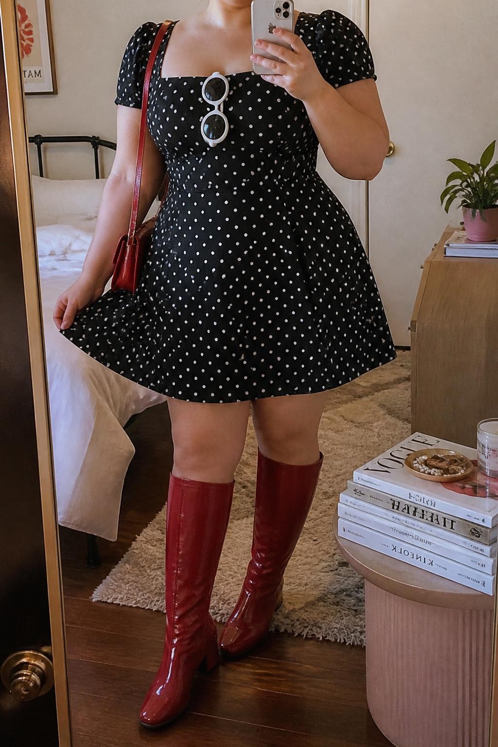 Mirror selfie of a plus size woman wearing a black and white polka-dot A-line mini dress with short puff sleeves and a fitted waist, cherry red knee-high boots with a low block heel, a small cherry red shoulder bag, and white oval sunglasses, clearly showing the exact dress print, colors, sleeve shape, footwear, and accessories listed, holding a phone in front of a mirror, cropped at the neck so her face does not show, relaxed playful selfie pose, in a cozy bedroom mirror setup with soft indoor lighting, a stack of fashion magazines on a side table and a small plant on a dresser, realistic retro festival styling, latest iPhone photo quality.