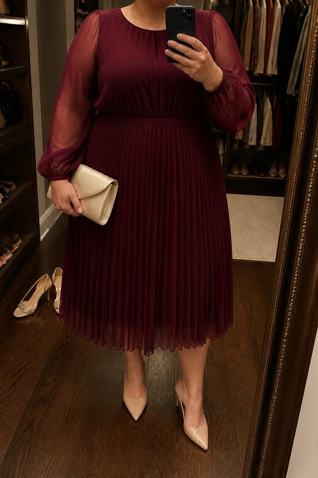 Mirror selfie of a plus size woman wearing a burgundy chiffon pleated midi dress with long sheer sleeves, a lined bodice, and a softly gathered waist, nude pointed pumps with low heels, a champagne satin clutch, and small crystal stud earrings, clearly showing the exact clothing items, burgundy chiffon, pleats, sheer sleeves, colors, footwear, and accessories listed, holding a phone in front of a mirror, cropped at the neck so her face does not appear, relaxed polished selfie stance, in a walk-in closet mirror with warm moody lighting, an open closet with clothes visible and a pair of heels on the floor nearby, realistic formal wedding styling, latest iPhone photo quality.
