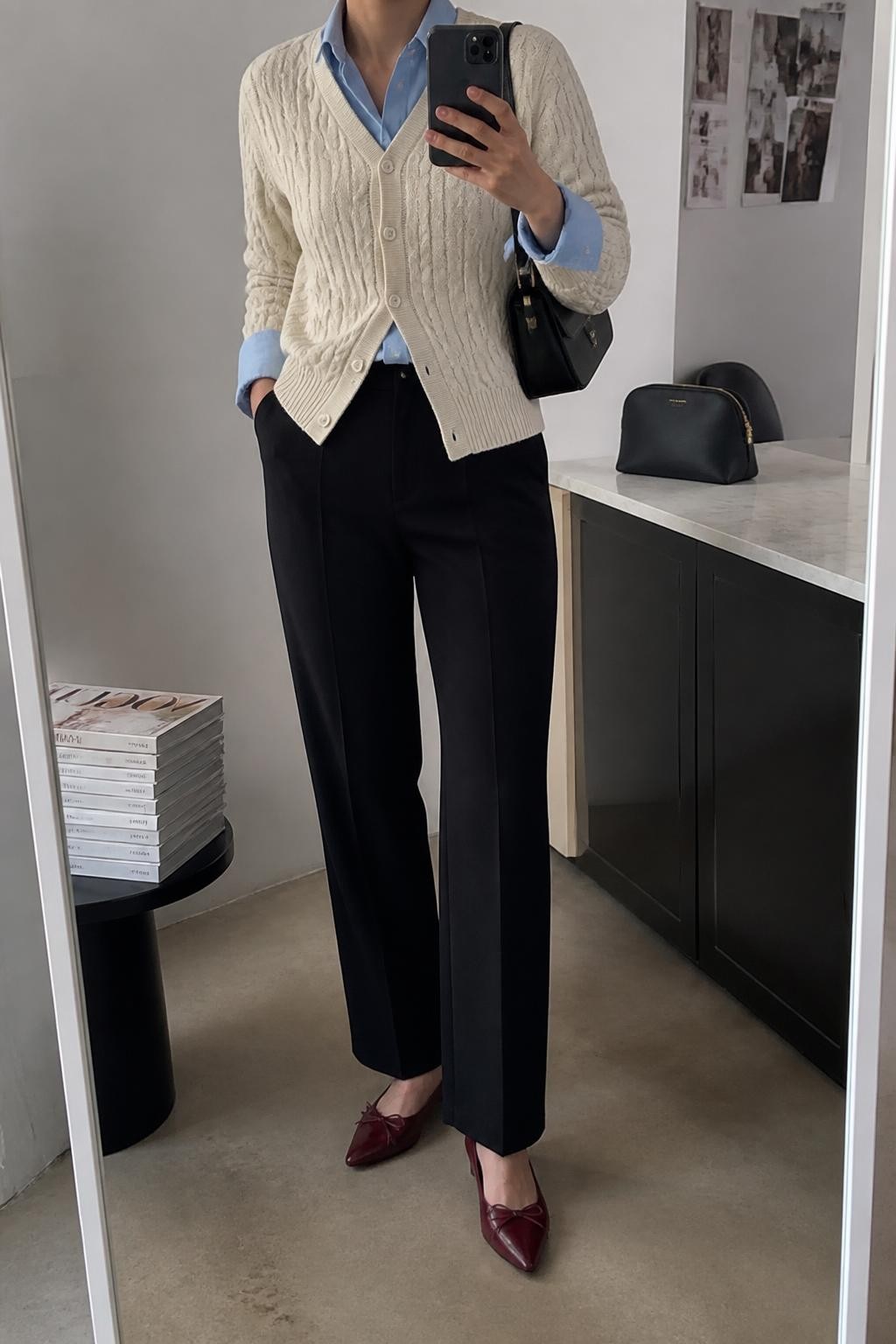 mirror selfie of a woman wearing a cream V-neck cable-knit cardigan with the bottom buttons undone over a pale blue cotton button-down shirt with the collar and cuffs visible, black high-waisted straight-leg trousers, burgundy pointed-toe ballet flats, and a black structured shoulder bag, clearly showing the exact clothing items, colors, fabrics, shoes, and bag listed in the Outfit Pieces section, holding a phone in front of a mirror, cropped at NECK so the face does not show, relaxed professional selfie pose in a fashion studio mirror corner, soft indoor lighting, with a stack of fashion magazines on a side table and a cosmetic bag on the counter, realistic cable knit, cotton, trouser, and leather textures, latest iPhone photo quality