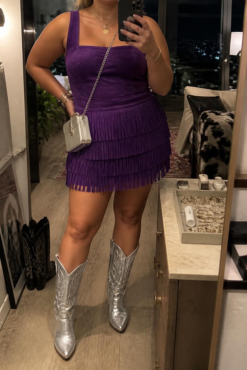 Mirror selfie of a plus size woman wearing a purple suede mini dress with wide straps, a square neckline, a fitted waist, and layered fringe across the skirt, metallic silver mid-calf cowboy boots with stitched details, and a small silver box crossbody bag, clearly showing the exact clothing items, purple suede, layered fringe, silver footwear, and accessories listed, holding a phone in front of a mirror, cropped at the neck so the face does not show, relaxed energetic selfie pose, in a stylish city apartment with warm moody lighting, a jewelry tray on the dresser and a pair of boots on the floor nearby, realistic evening Coachella outfit, latest iPhone photo quality.