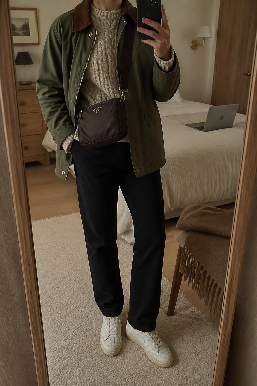mirror selfie of a woman wearing an open olive cotton barn jacket with a brown corduroy collar over an oatmeal cable-knit crewneck sweater, black straight-leg stretch travel trousers, cream leather sneakers, and a chocolate nylon crossbody bag resting near the waist, clearly showing the exact clothing items, colors, fabrics, sneakers, and bag listed in the Outfit Pieces section, holding a phone in front of a mirror, cropped at NECK so the face does not show, relaxed casual selfie pose in a cozy bedroom mirror setup, warm moody lighting, with a laptop on the bed and a folded blanket on a chair, realistic cotton, corduroy, cable knit, stretch fabric, leather, and nylon textures, latest iPhone photo quality
