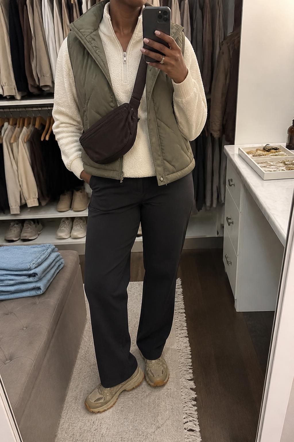 mirror selfie of a Black woman wearing an open olive quilted puffer vest over a cream half-zip fleece pullover, black straight-leg hiking pants, tan trail sneakers with textured soles, and a chocolate nylon belt bag worn crossbody, clearly showing the exact clothing items, colors, fabrics, sneakers, and bag listed in the Outfit Pieces section, holding a phone in front of a mirror, cropped at NECK so the face does not show, relaxed outdoor-ready selfie pose in a walk-in closet mirror, soft indoor lighting, with folded denim jeans on a bench and a jewelry tray on the vanity, realistic quilting, fleece, technical fabric, rubber, and nylon textures, latest iPhone photo quality