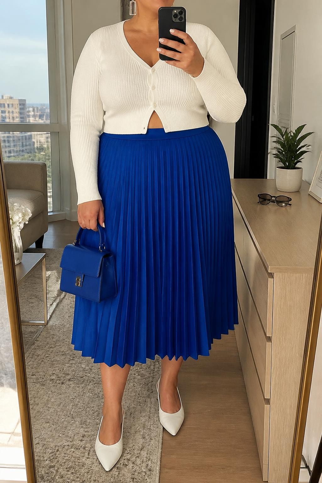 Mirror selfie of a plus size woman wearing a cobalt blue high-waisted pleated midi skirt, a fitted white cropped cardigan buttoned at the front, white pointed slingback flats, a structured cobalt mini handbag, and silver rectangular earrings, clearly showing the exact clothing items, cobalt pleats, white knit, colors, footwear, and accessories listed, holding a phone in front of a mirror, cropped at the neck so the face does not show, relaxed bright selfie pose, in a stylish city apartment with natural window light, sunglasses placed on a dresser and a small plant nearby, realistic spring outfit, latest iPhone photo quality.