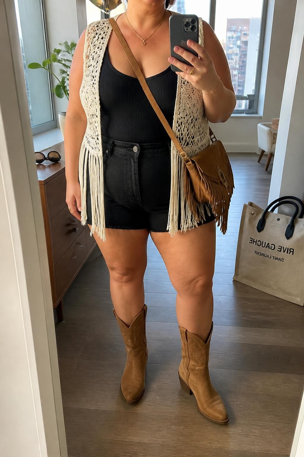 Mirror selfie of a plus size woman wearing a cream crochet fringe vest over a fitted black ribbed scoop-neck bodysuit, black high-waisted denim shorts with relaxed thigh openings, tan leather mid-calf cowboy boots, and a small tan suede fringe crossbody bag, clearly showing the exact clothing items, cream crochet texture, black denim, tan footwear, and accessories listed, holding a phone in front of a mirror, cropped at the neck so the face does not show, relaxed festival selfie pose, in a stylish city apartment with natural window light, sunglasses placed on a dresser and a tote bag resting on the floor, natural unstaged Coachella outfit, latest iPhone photo quality.