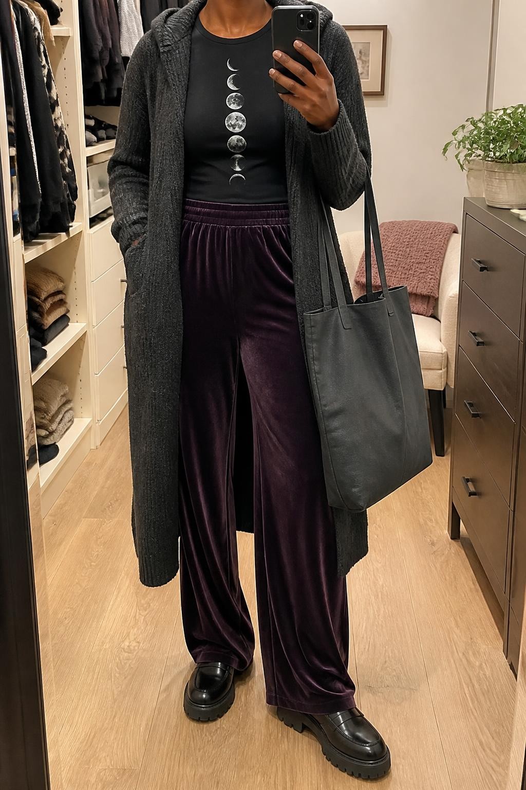 mirror selfie of a Black woman wearing an open charcoal longline hooded knit cardigan over a black fitted top with a clear silver moon-phase print tucked into plum wide-leg velvet trousers, black chunky leather loafers, and a plain charcoal canvas tote with no writing, clearly showing the exact clothing items, colors, print, fabrics, shoes, and bag listed in the Outfit Pieces section, holding a phone in front of a mirror, cropped at NECK so the face does not show, relaxed cozy selfie pose in a walk-in closet mirror, soft indoor lighting, with a folded blanket on a chair and a small plant on a dresser, realistic knit, printed fabric, velvet, leather, and canvas textures, natural unstaged mirror photo, latest iPhone photo quality