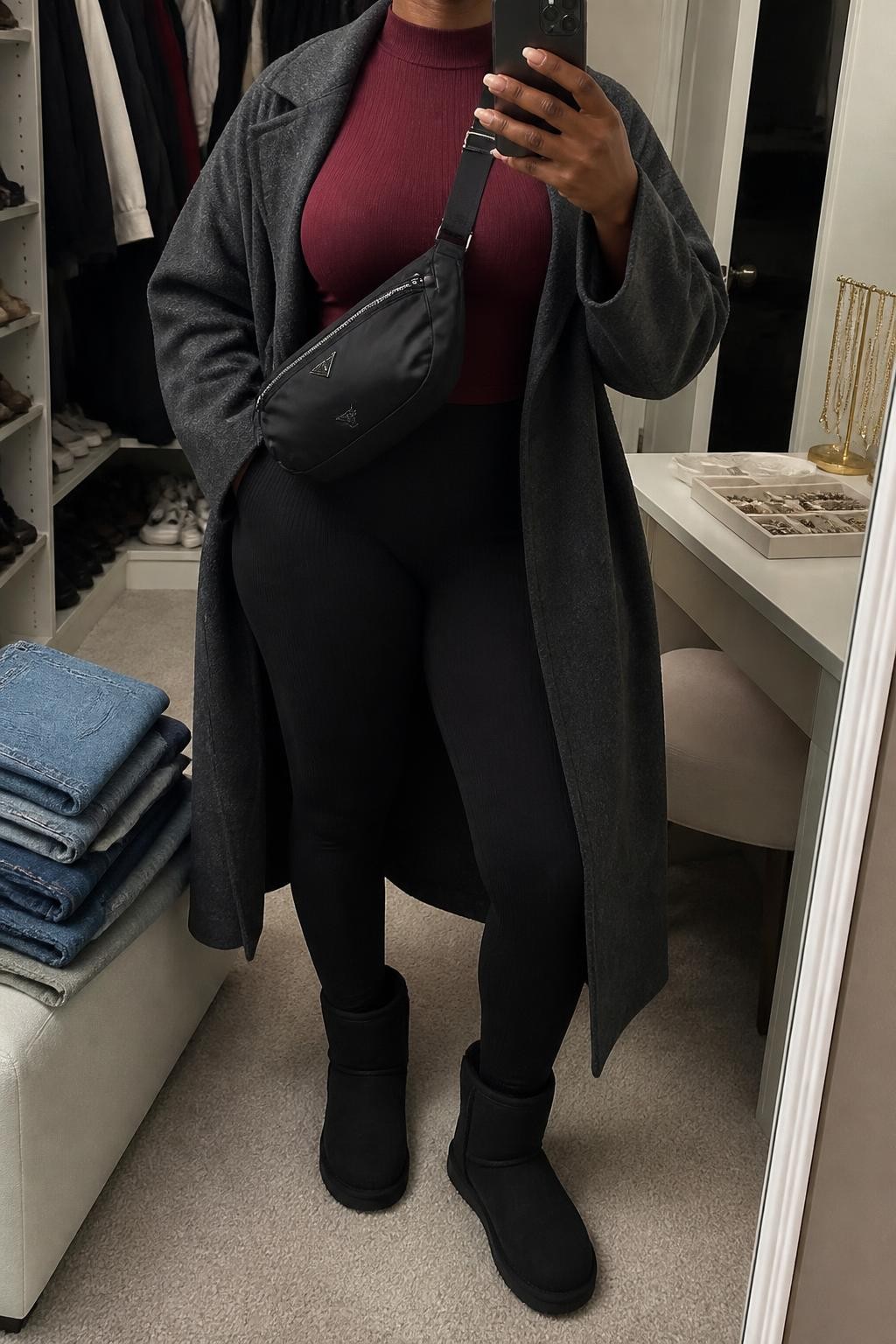 mirror selfie of a Black woman wearing an open charcoal gray oversized wool-blend coat over a burgundy fitted ribbed mock-neck top, black high-waisted ribbed leggings, black ultra mini Ugg boots, and a black nylon crossbody bag resting near the waist, clearly showing the exact clothing items, colors, fabrics, boots, and bag listed in the Outfit Pieces section, holding a phone in front of a mirror, cropped at NECK so the face does not show, relaxed casual selfie pose in a walk-in closet mirror, soft indoor lighting, with folded denim jeans on a bench and a jewelry tray on the vanity, realistic wool, ribbed knit, nylon, and suede textures, latest iPhone photo quality