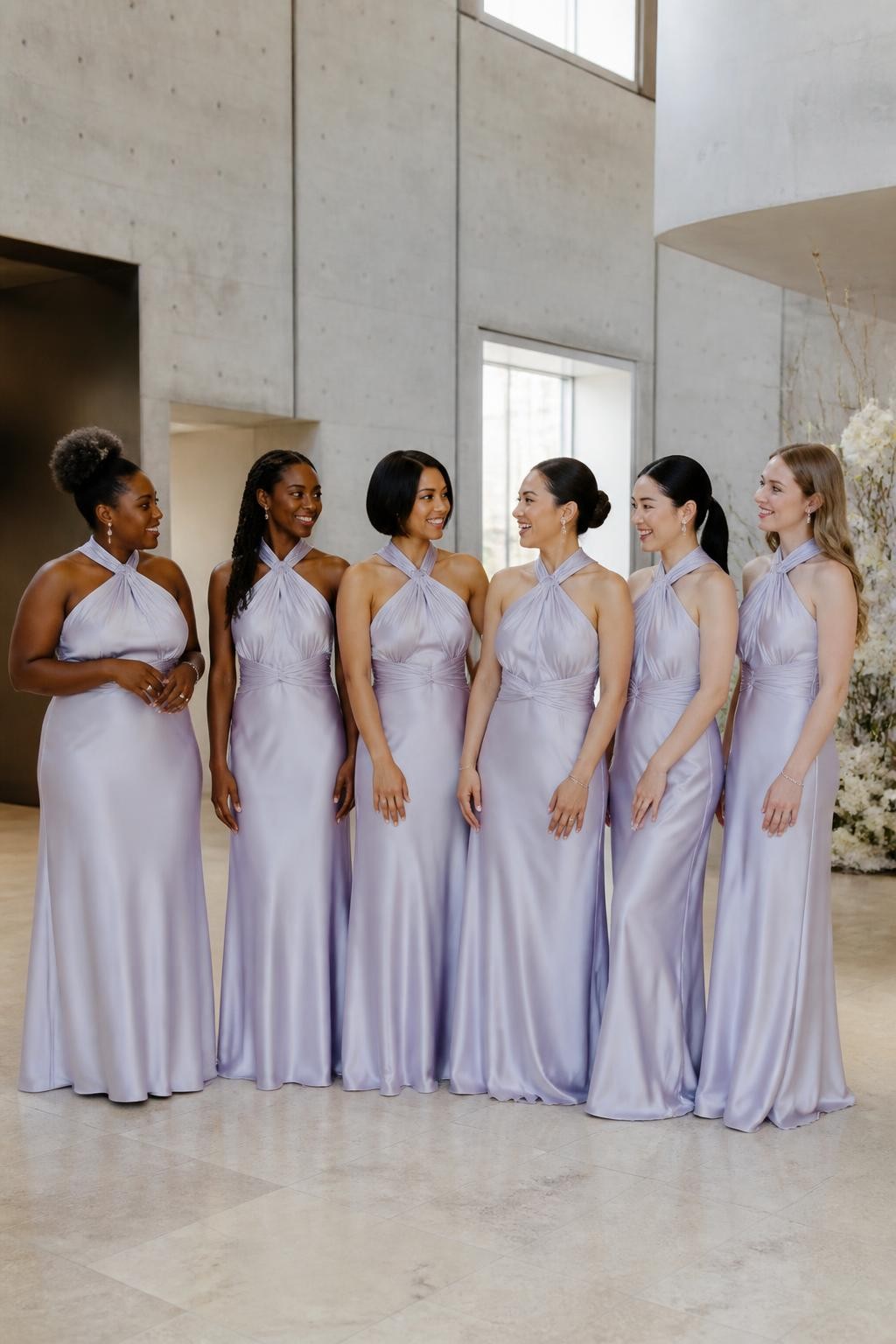 Realistic contemporary wedding photograph of six bridesmaids wearing icy lavender satin gowns with twisted halter necklines, soft folds wrapping around the neck, open upper backs, lightly gathered waists, and fluid floor-length skirts, women with deep blue-black, dark warm brown, copper, golden olive, East Asian fair, and pale rosy complexions, different shoulder widths, torso lengths, ages, and body shapes represented realistically, bright confident faces with individual features and subtle makeup, hairstyles including a high curly puff, long twists, polished side-part bob, sculptural bun, sleek ponytail, and brushed waves, standing inside a modern art museum with pale concrete walls, limestone floors, tall windows, and minimal white floral installations, directional daylight revealing the cool silver-lavender undertone, realistic satin creasing, natural posture, and relaxed group spacing.